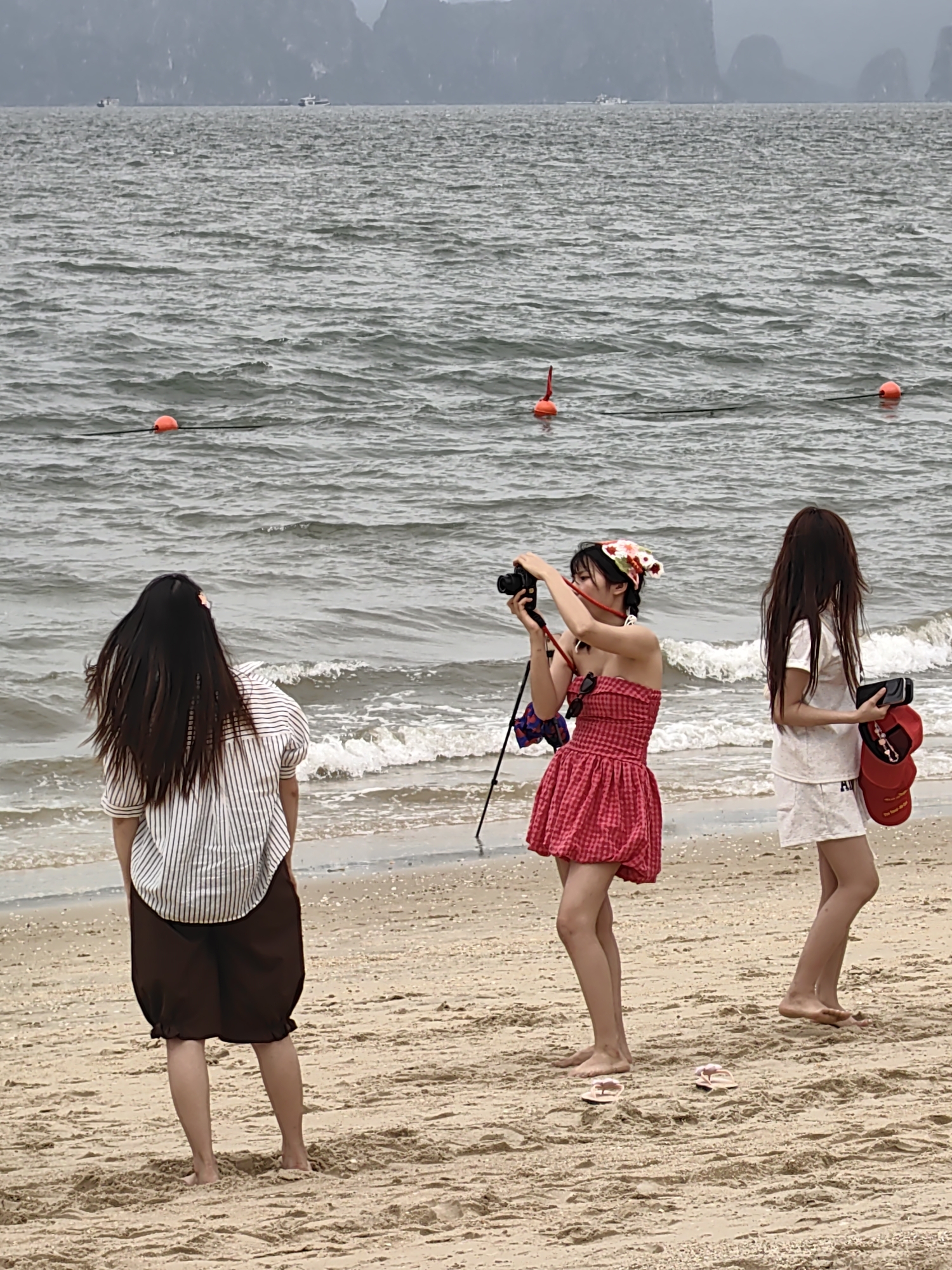 The image shows three young women on a sandy beach with a choppy body of water in the background, marked with several orange buoys. The background also features distant, hazy hills, likely rock formations, in Bai Chay Ward, Vietnam. The woman in the center is taking a photograph with a black camera. She is wearing a red and white checkered dress with a colorful bandana on her head. A camera tripod is visible behind her. The woman to the left is facing away from the camera, wearing a striped shirt and brown shorts. The woman on the right is wearing a white t-shirt and shorts, holding a phone and a red hat. All three women are barefoot. The sand is light brown.