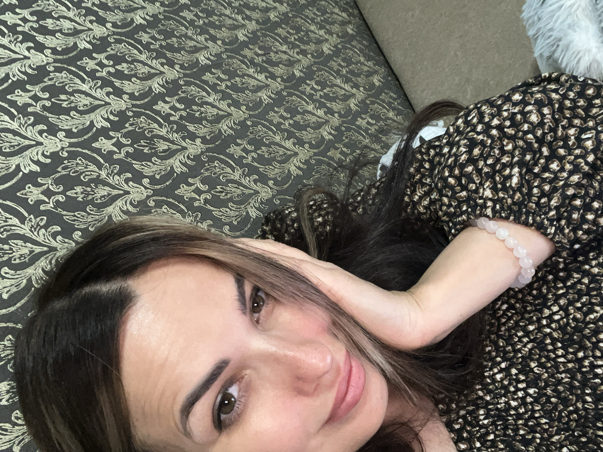 A woman is captured in a close-up selfie, her head tilted slightly as she rests her right hand against her cheek. She has long, dark brown hair with lighter highlights, which falls across her face. Her eyes are brown, and she has a subtle smile. She is wearing a patterned top with black and brown tones, and a bracelet made of light pink beads is visible on her wrist. The background is a wall with a textured, ornate pattern in shades of dark gray and gold. The lighting suggests an indoor setting, possibly a room with soft illumination. No specific location details are discernible from the image itself.
