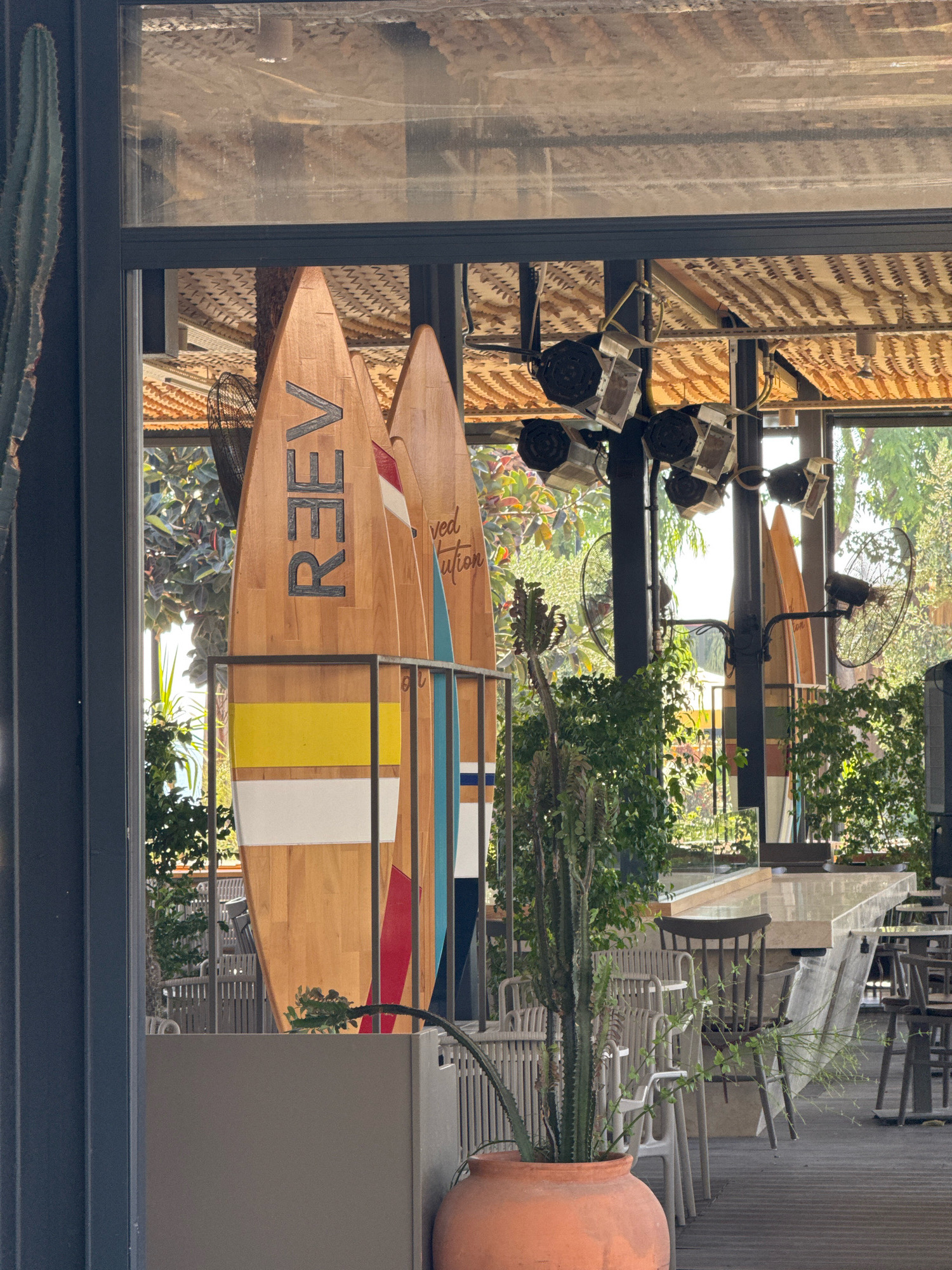 The image depicts the interior of a restaurant or cafe, likely in Antalya, Turkey, with a bohemian or beach-themed ambiance. Large wooden surfboards, some with painted designs and lettering like "RE3EV," are prominently displayed as decorative elements, leaning against a wall or partition. Behind the surfboards, large windows offer a view of an outdoor seating area with tables and chairs, surrounded by lush greenery and trees. The interior ceiling is adorned with woven natural materials and features industrial-style lighting fixtures and fans. Several potted plants, including a tall cactus in an terracotta pot, are placed throughout the seating area, enhancing the tropical or relaxed atmosphere. The overall scene suggests a casual, open-air dining environment, possibly a popular spot for tourists and locals alike, given Antalya's coastal location and appeal.