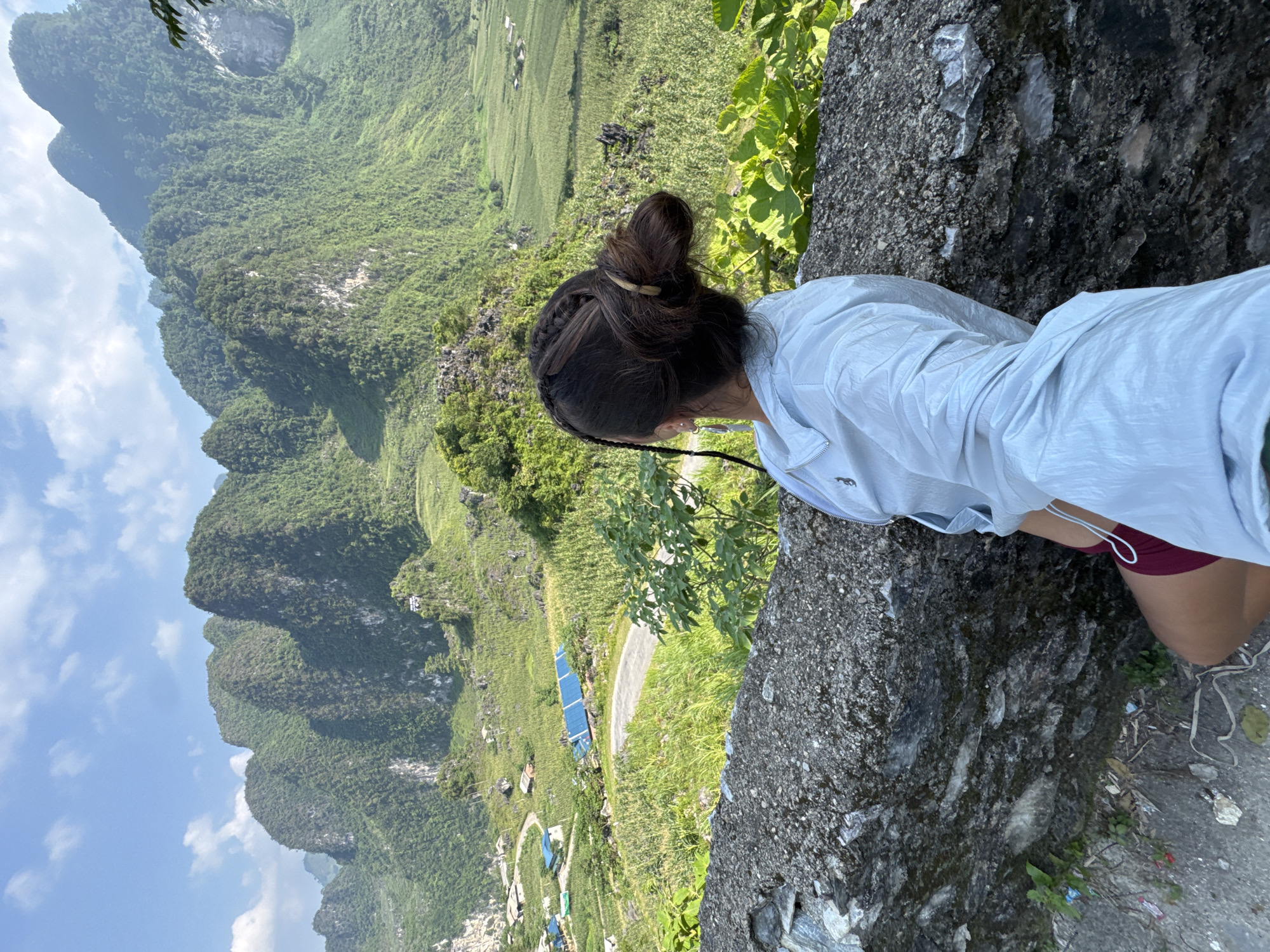 A person with their hair in a bun, wearing a light blue jacket and maroon shorts, is seen from behind, looking over a rocky ledge. The landscape below features lush, green mountains under a partly cloudy sky, and a road with buildings and fields in Đường Thượng Commune, Vietnam.