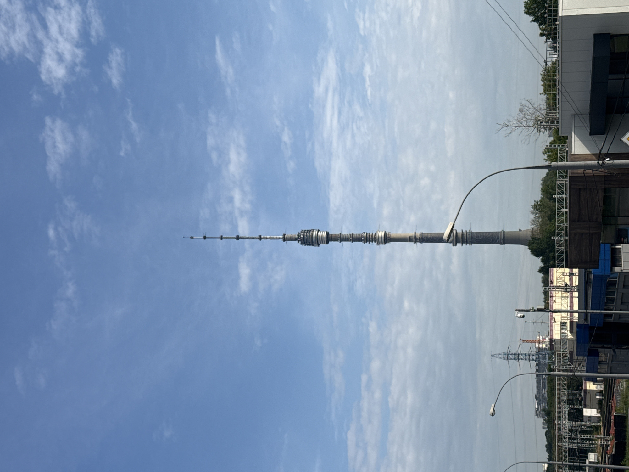 The image shows the Ostankino Tower, a prominent landmark in Moscow, Russia, dominating the skyline under a partly cloudy blue sky. The tower is a tall, slender telecommunications and broadcasting tower, with a distinct segmented structure. In the foreground, there are various urban elements, including train tracks, industrial buildings, streetlights, and power lines. The overall scene depicts an industrial or commercial area of Moscow with the iconic tower as the central focus. There are no people visible in the image.