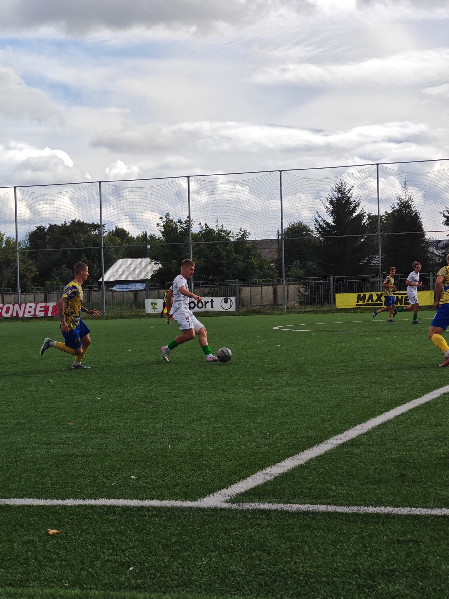 A football match is taking place on a green turf field under a cloudy sky. Three male athletes are visible on the field. In the foreground, one player in a white jersey with the number 7 and green socks is kicking a black and white football with his right foot. To his left and slightly behind him, a player in a yellow and blue jersey with a patterned design is running. Further in the background, another player in a white jersey is running on the field. On the left side of the frame, a banner with "FONBET" written on it is visible. On the right side, a yellow banner with "MAXINE" is displayed. A chain-link fence with a net runs along the length of the field. Trees and buildings are visible in the background behind the fence. The scene appears to be at a sports complex in Homyel, Belarus.