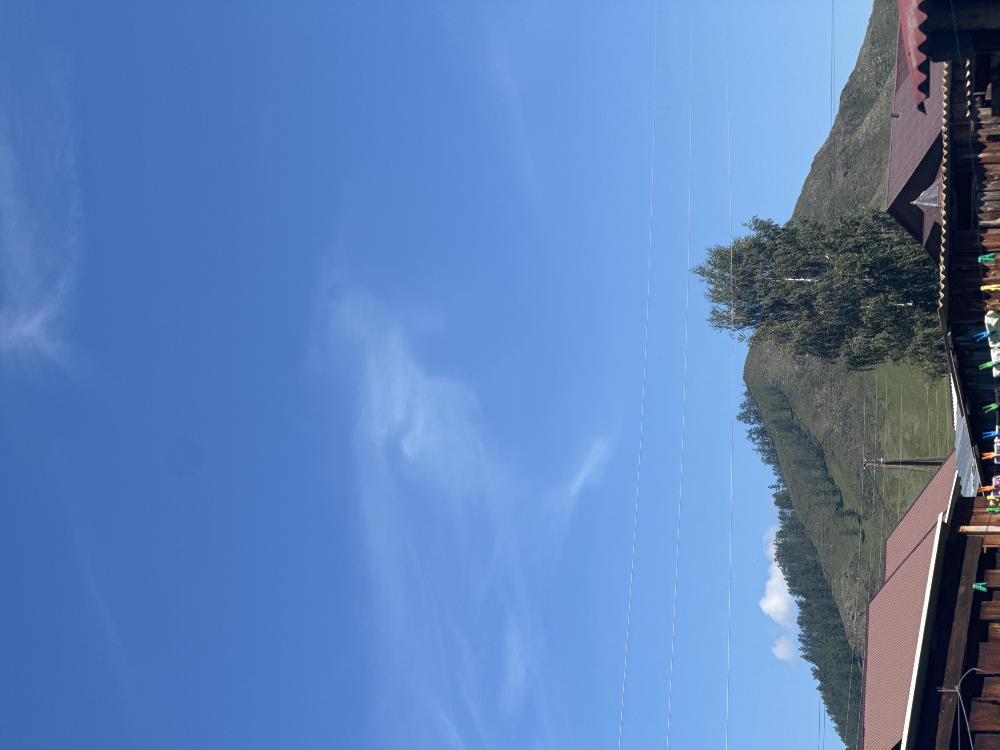 This is a full-frame, eye-level shot of a rural landscape in Yabogan, Russia, on a clear, sunny day. The image is dominated by a bright blue sky with thin, wispy clouds. In the mid-ground, a gently sloping, treed mountain rises, its green slopes dotted with dark green conifers. In the foreground, the roofs of several wooden structures, likely houses or sheds, are visible. These buildings have reddish-brown, corrugated metal roofs. A tall, leafy tree with a white trunk stands to the right of the mountain. Clotheslines with clothes clipped to them are strung across the foreground, suggesting a domestic setting. Several power lines stretch horizontally across the image.