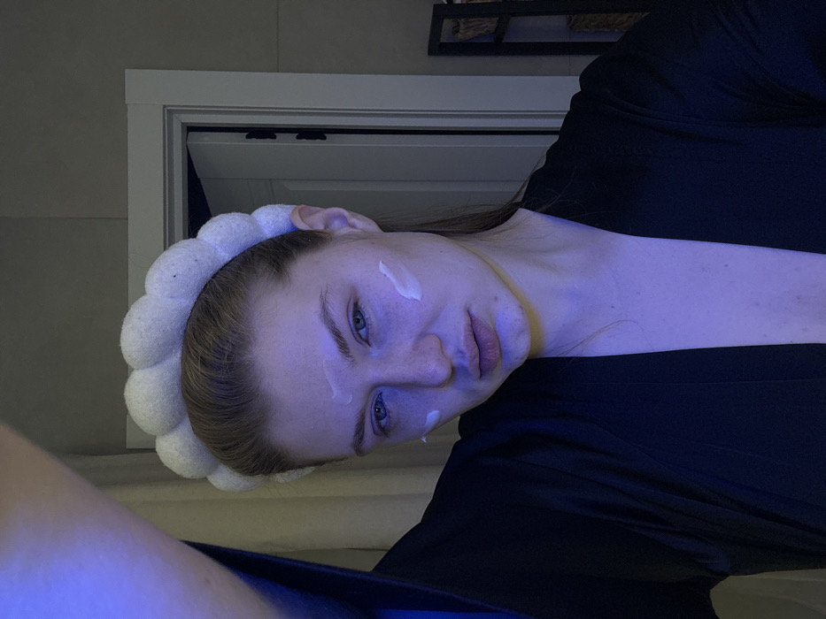 A person with fair skin is shown from a close, tilted angle. They wear a white, puffy headband and have dabs of cream on their face. They are wearing a black garment. The lighting has a cool, blue tone. Behind the person is a white door frame and possibly a shelf with decorations on the wall behind. The picture appears to be taken indoors.