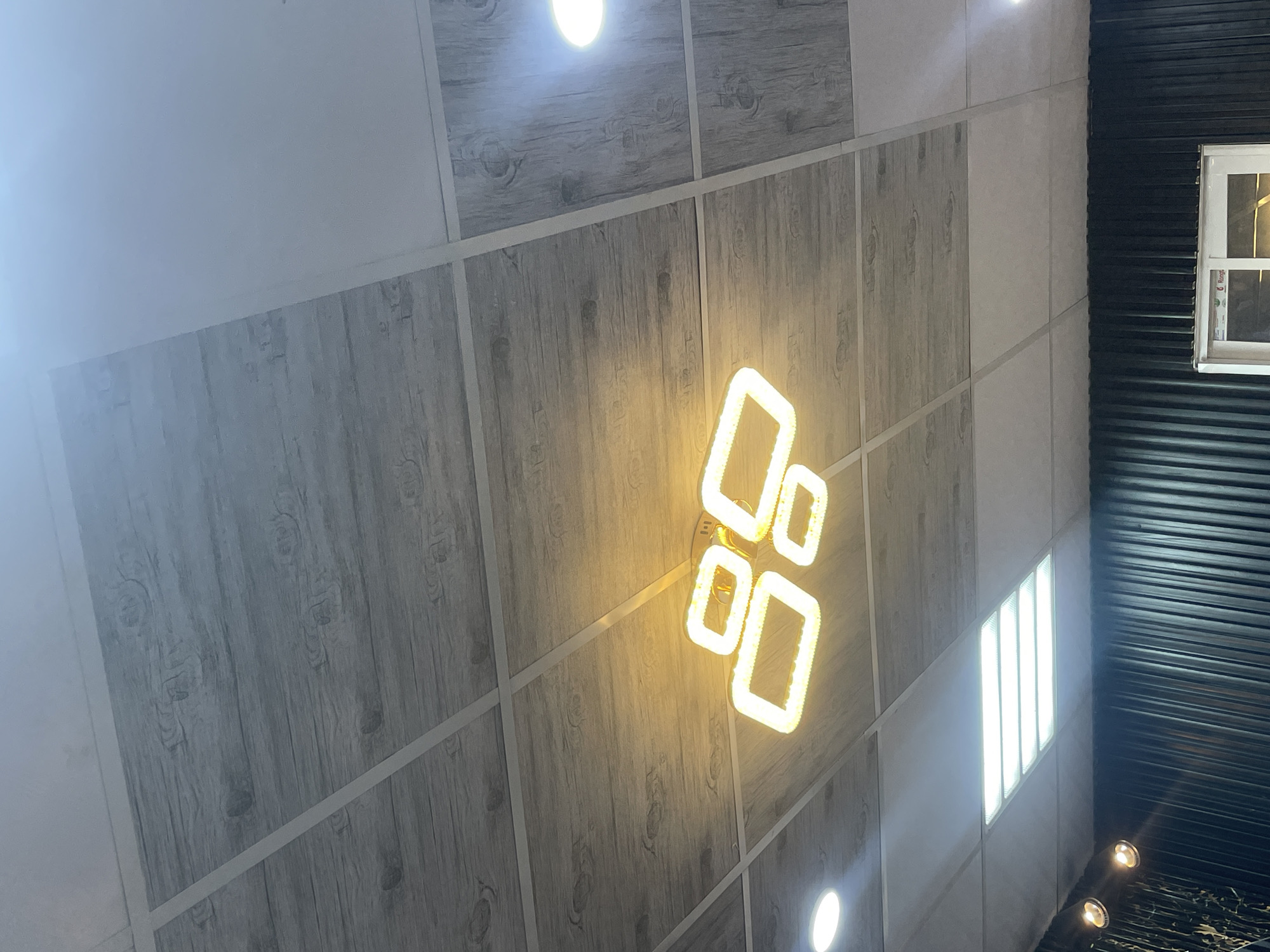 The image shows a ceiling with wood-patterned tiles and white panels. A modern, rectangular chandelier with multiple illuminated rectangular frames hangs from the ceiling. Two round ceiling lights are also visible. Below the ceiling, a black corrugated wall is partially visible, with a white-framed window. The scene appears to be an interior space in Barako, Nigeria, likely a commercial or public area, given the ceiling design and lighting fixtures. No people or specific actions are depicted. The lighting is warm from the chandelier and cool from the ceiling lights.
