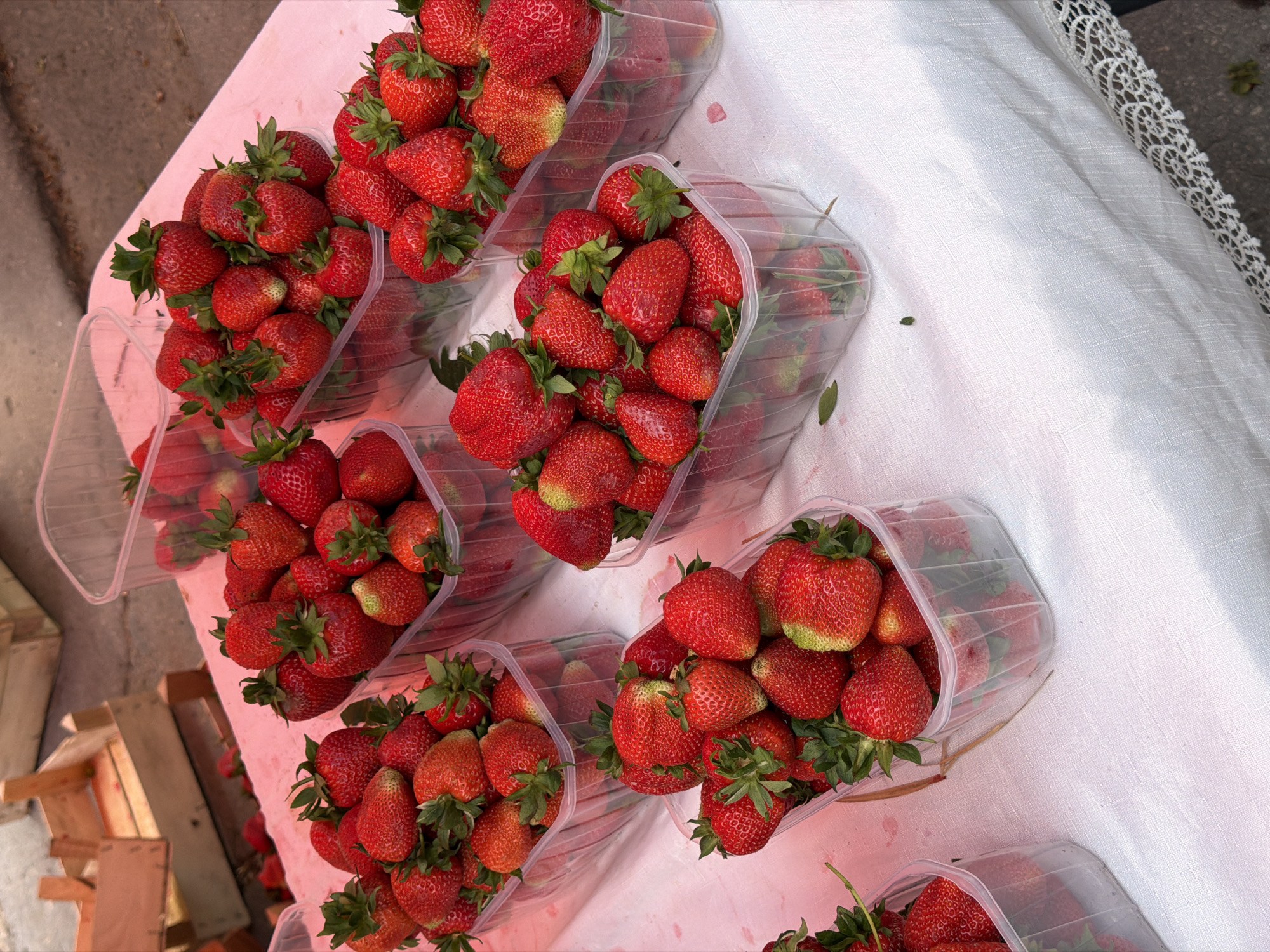 A close-up, high-angle shot captures a market stall overflowing with fresh strawberries. Several clear plastic containers are filled to the brim with ripe, red strawberries, their green stems and leaves adding a touch of contrast. The containers are arranged on a white tablecloth, which is partially covered by a pink surface, possibly a market stall covering. In the background, wooden crates are visible, suggesting a retail setting. The scene is set outdoors, with a glimpse of an asphalt surface in the lower right corner. The image was taken at the Central Market in Chișinău, Moldova.