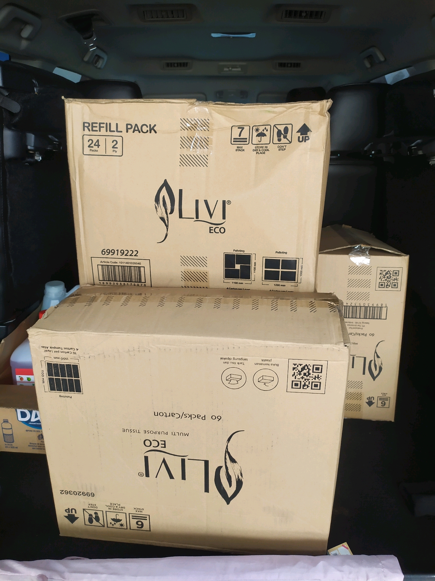 This image depicts the interior of a vehicle, likely an SUV, filled with several large cardboard boxes of paper products. The boxes are stacked and arranged within the cargo area of the vehicle.
The most prominent box, in the foreground, is labeled "Livi ECO MULTI PURPOSE TISSUE 60 Packs/Carton" and features a QR code and various icons indicating product handling instructions. Another box behind it, also labeled "Livi ECO," is partially visible. A third, larger box in the background is labeled "REFILL PACK 24 Packs 2 Ply" and also shows "Livi ECO" branding, along with handling icons. Other smaller boxes and items, including what appears to be a bottle of cleaning solution and a clear plastic bottle, are also present. The overall impression is of a delivery or storage of bulk paper goods. The presence of the vehicle's interior, including the ceiling and seats, suggests an indoor or covered parking location, possibly at a distribution center or a residence in Sleman, Indonesia.