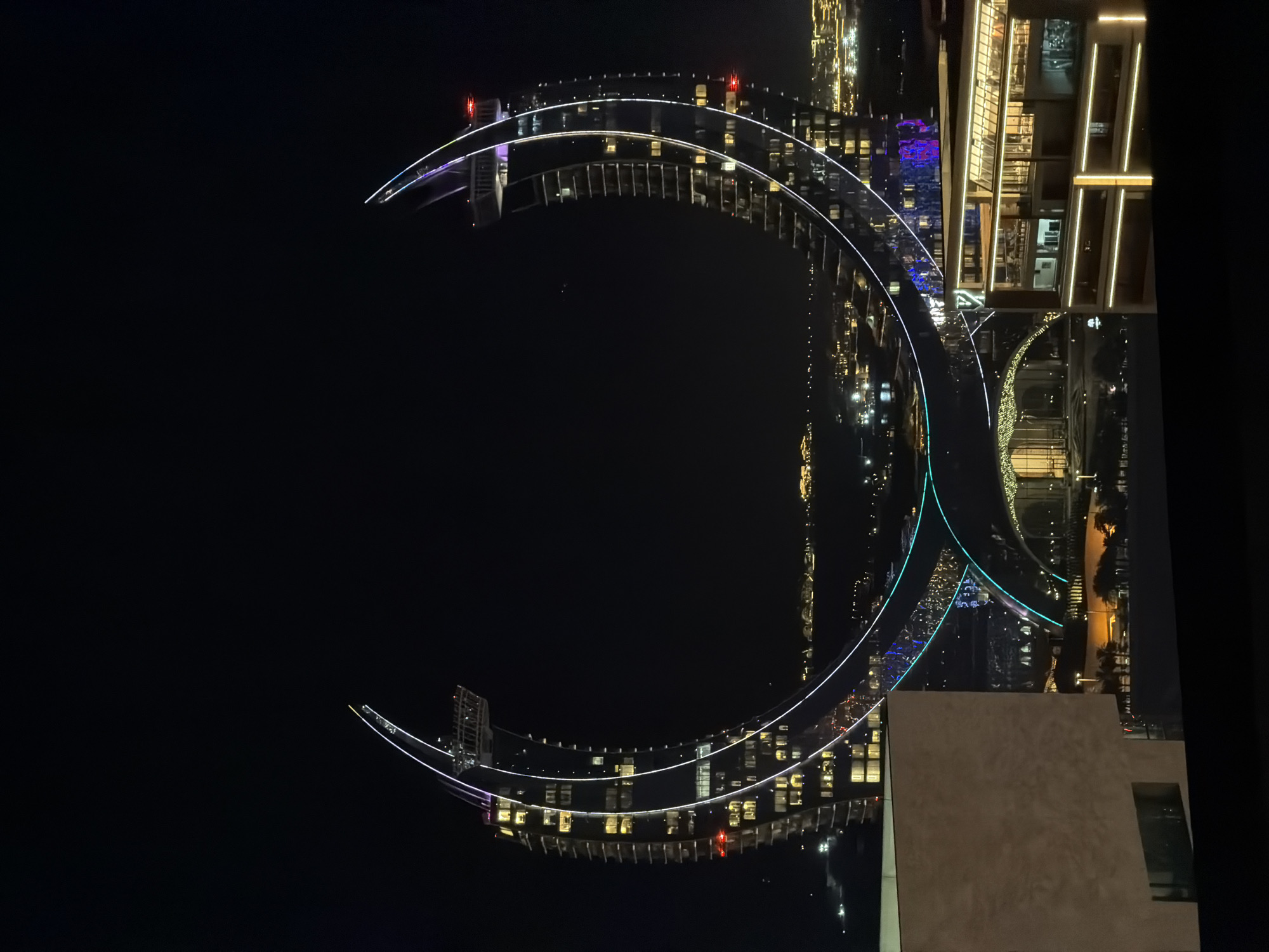 An eye-level, exterior, low-angle night shot captures the crescent-shaped architecture of the Marsa Malaz Kempinski hotel in Doha, Qatar. The hotel, illuminated with white and colored lights, is situated in a cityscape that extends to the horizon. The dark sky serves as a backdrop to the brightly lit structure. In the foreground, the corner of a tan, modern building is visible on the left, with a dark foreground of an unknown structure. The hotel's distinctive design features a curved facade with multiple floors, each adorned with numerous illuminated windows. Two curving, illuminated walkways or bridges connect the two towers of the hotel, adding to its dramatic nighttime appearance. The cityscape in the background is a collection of illuminated buildings and structures, suggesting a well-developed urban environment.