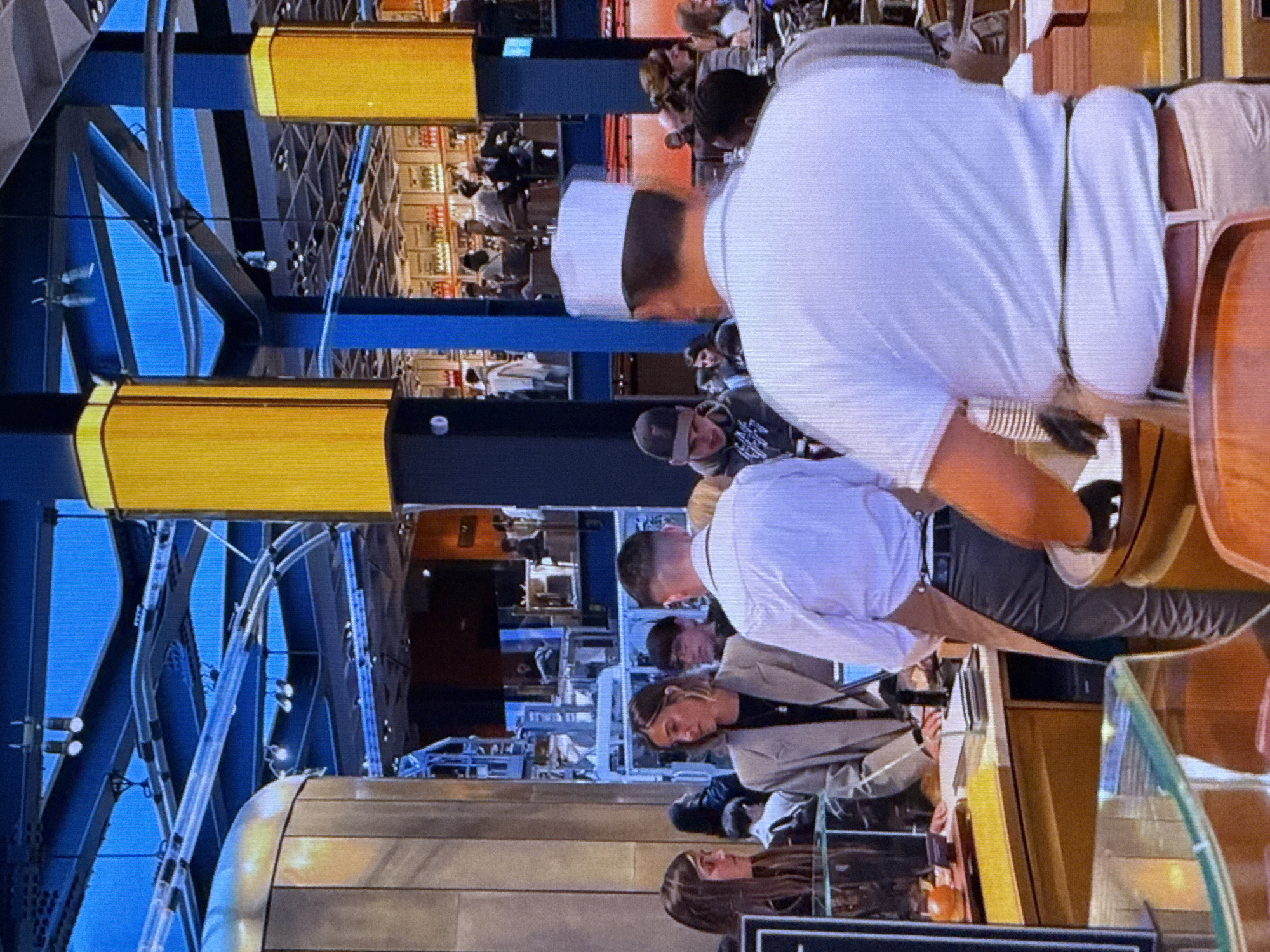 The image depicts an indoor scene at what appears to be a busy retail or food establishment, possibly a market or a restaurant with multiple food stalls. The setting features a modern architectural design with prominent blue and yellow structures, possibly shelving or decorative elements. Metal beams and conduits are visible throughout the space, suggesting an industrial or functional aesthetic.
Several individuals are present, engaged in various activities. In the foreground, a person wearing a white chef's hat and a white uniform is partially visible, leaning over a counter. Behind them, other people are standing and interacting. Some individuals appear to be customers, while others might be staff. One person in the mid-ground is wearing a light-colored jacket, and another is wearing a dark beanie. The general atmosphere seems active and bustling.
The lighting suggests it's daytime, but there are no windows visible to confirm natural light or weather conditions. The overall impression is of a vibrant commercial environment. There is no visible text in the image. The presence of food stalls and people interacting with them indicates that a transaction or food service is likely taking place. The perspective of the image is from a slightly elevated viewpoint, looking down into the scene.