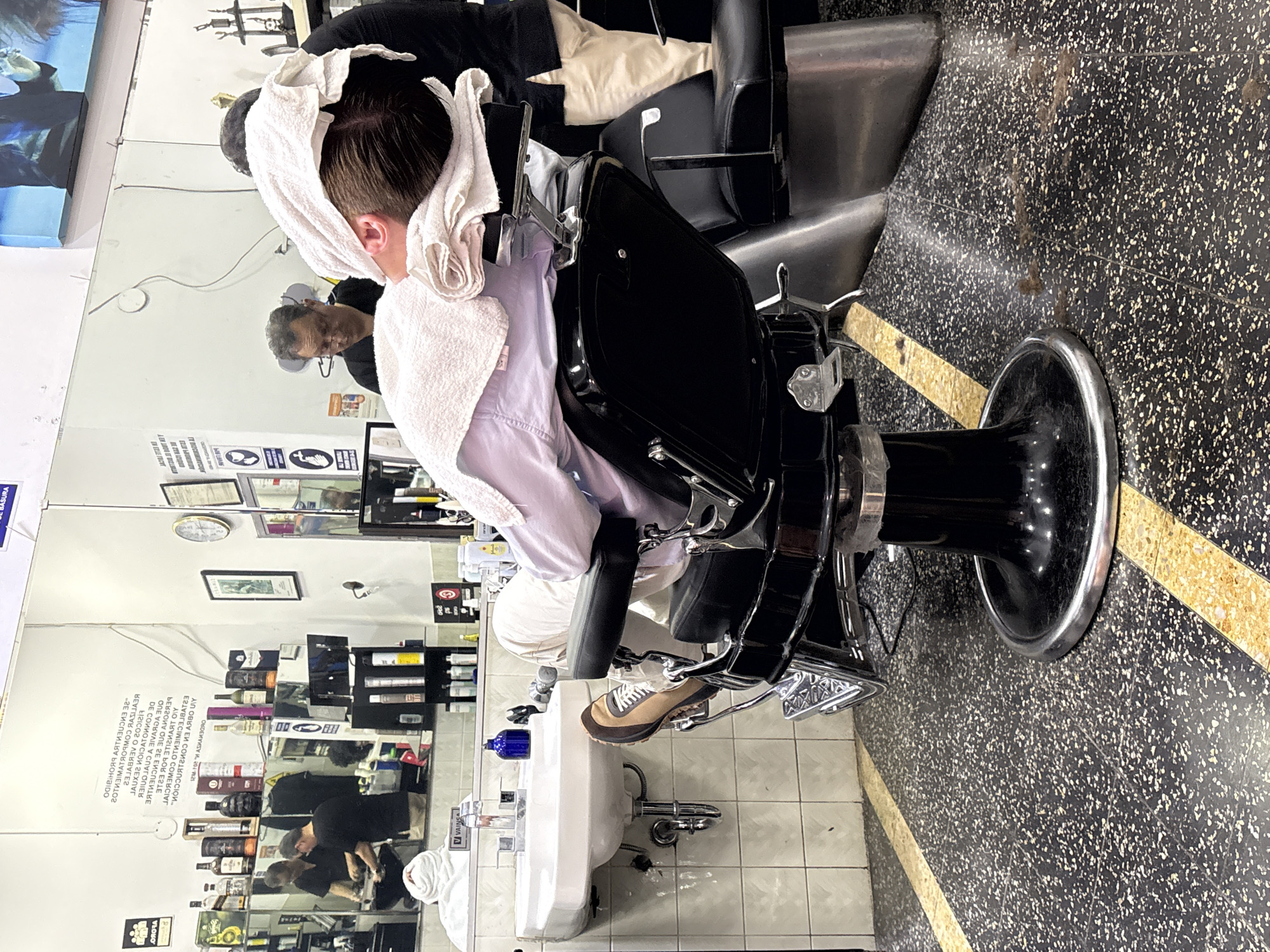 The image depicts the interior of a barber shop in San Isidro, Peru. A man is seated in a black barber chair, reclined with a white towel draped over his head and neck. He is wearing a light purple shirt and cream-colored pants, with his right leg resting on a footrest. The barber chair is ornate, with chrome accents and a black leather seat. In the background, a sink with a white basin is visible, along with shelves stocked with various bottles, presumably hair products. Another man, the barber, stands behind the seated customer, wearing glasses and a dark shirt. The floor is a speckled black and white terrazzo with yellow dividing lines. Mirrors line the wall behind the barber chair, reflecting shelves of products and other barber chairs. The overall atmosphere suggests a typical barber shop setting.