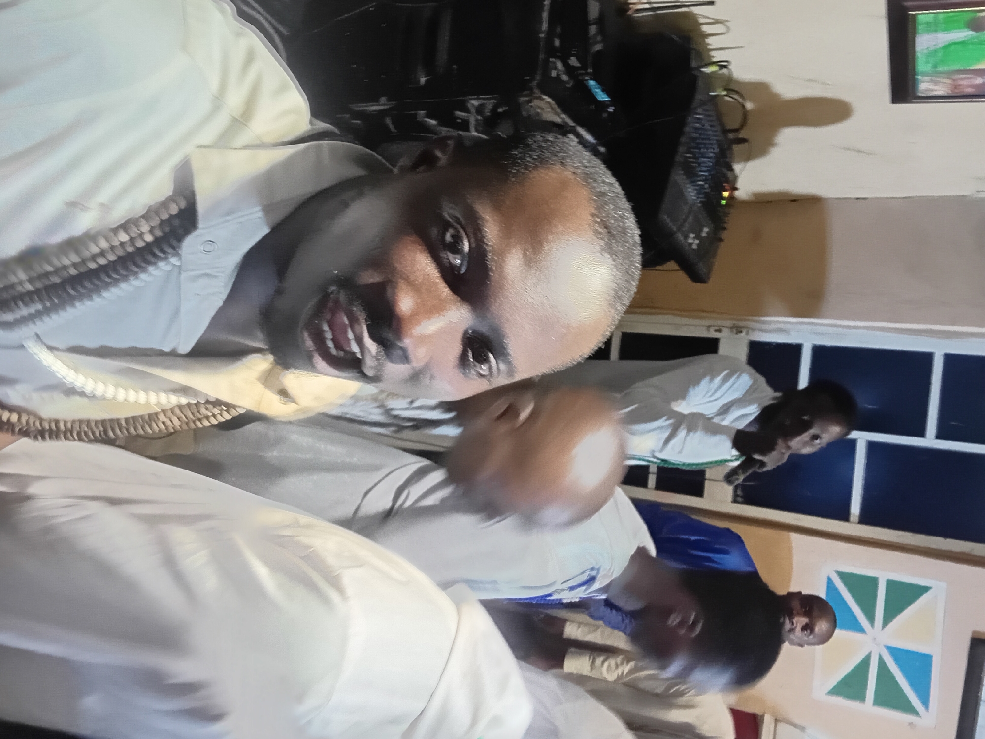In the city of Ajibo, Nigeria, the image captures a group of men indoors, possibly at an event or gathering. In the foreground, a man with dark skin and short hair is looking directly at the camera with his mouth open. He is wearing a light-colored collared shirt and a necklace made of what appears to be beads. Behind him, several other men are visible. One man in a white shirt is holding a microphone, suggesting he might be speaking or performing. Another man in a blue garment stands further back. In the background, a sound mixing board is present, indicating potential musical or audio equipment setup. A framed picture hangs on the wall to the left, and a decorative geometric design adorns the wall on the right. The environment appears to be a dimly lit indoor space.