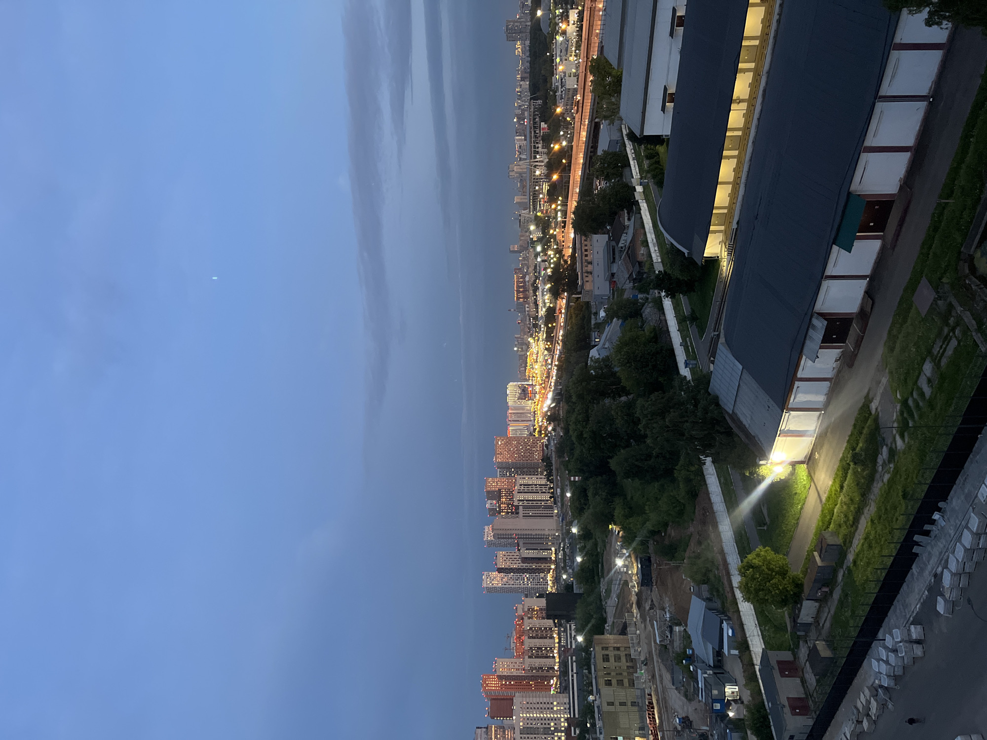 An elevated, eye-level, long shot captures a twilight cityscape, likely Tarasovka, Russia, with a clear, deep blue sky transitioning to darker hues near the horizon. A faint, vertical sliver of light, possibly a satellite or distant aircraft, is visible in the upper sky. The foreground features industrial buildings with dark, curved roofs and white facades, illuminated by bright, directional spotlights emanating from the left, casting long shadows and highlighting a grassy area. Adjacent to these are rows of concrete blocks and construction materials, suggesting ongoing development. A person is walking on a paved path in the lower left corner. Further into the mid-ground, a dense line of lush green trees separates the industrial structures from a sprawling urban landscape. This urban area is characterized by numerous high-rise residential buildings, many of which are illuminated with warm lights, creating a vibrant glow. A complex network of roads and highways, brightly lit by streetlights and vehicle headlights, dissects the cityscape, indicating a busy thoroughfare. In the distance, more buildings and structures are visible, fading into the hazy horizon. The overall scene depicts a transition from industrial outskirts to a densely populated urban center at dusk.