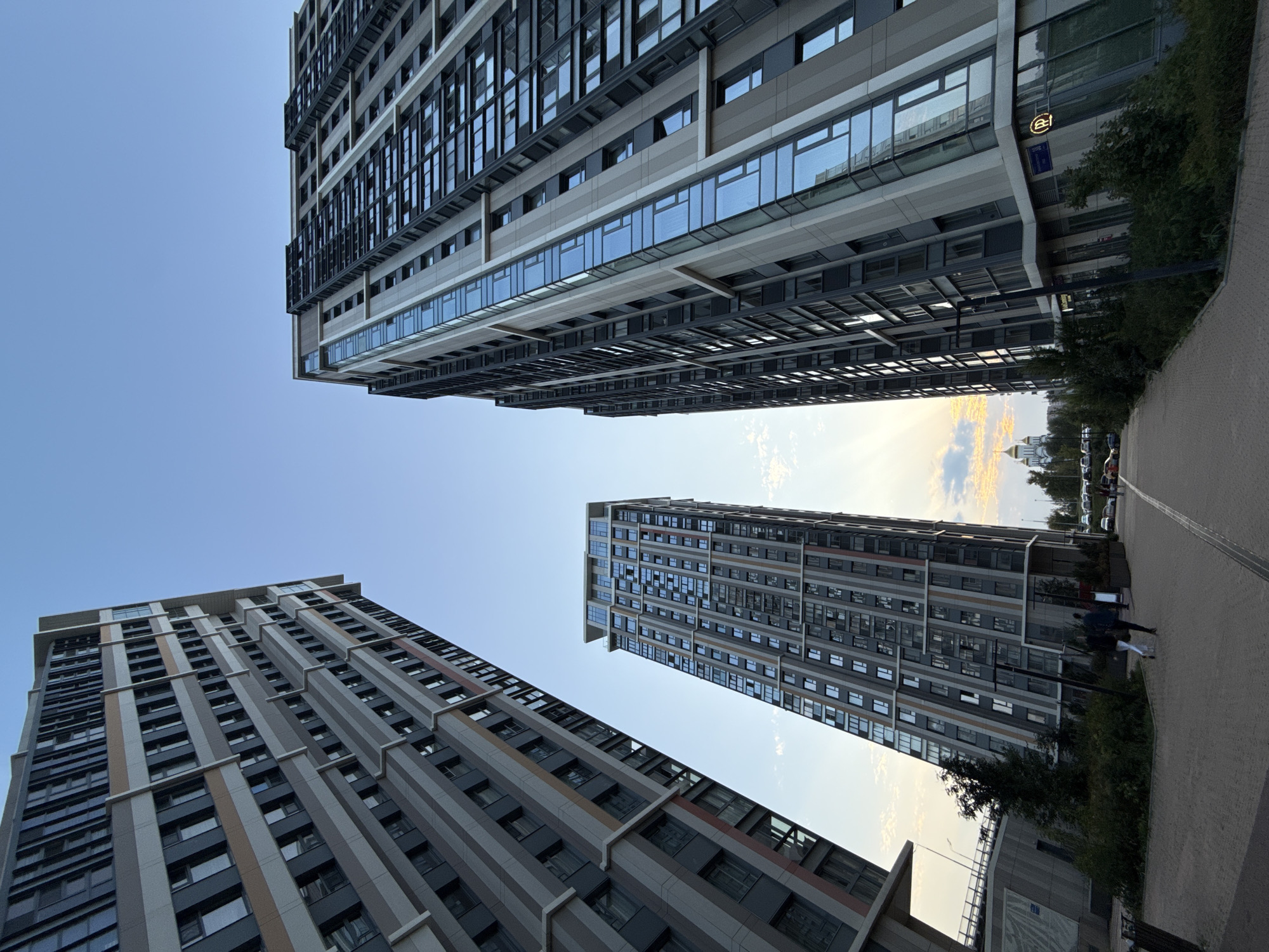 The image captures a low-angle view of modern high-rise apartment buildings against a clear blue sky. The buildings are characterized by their geometric facades with alternating bands of gray and beige, interspersed with numerous windows. On the left, a particularly tall building dominates the frame, angled upwards. To the right, another similar building stands, featuring more extensive glass panels. In the distance, a golden-domed building, likely a church, is visible, hinting at the location. A paved pathway leads towards the buildings, with some greenery and trees lining the sides. Two individuals are seen walking on the path. The overall scene suggests a contemporary urban residential area. This image was taken in the city of Moscow, Russia.