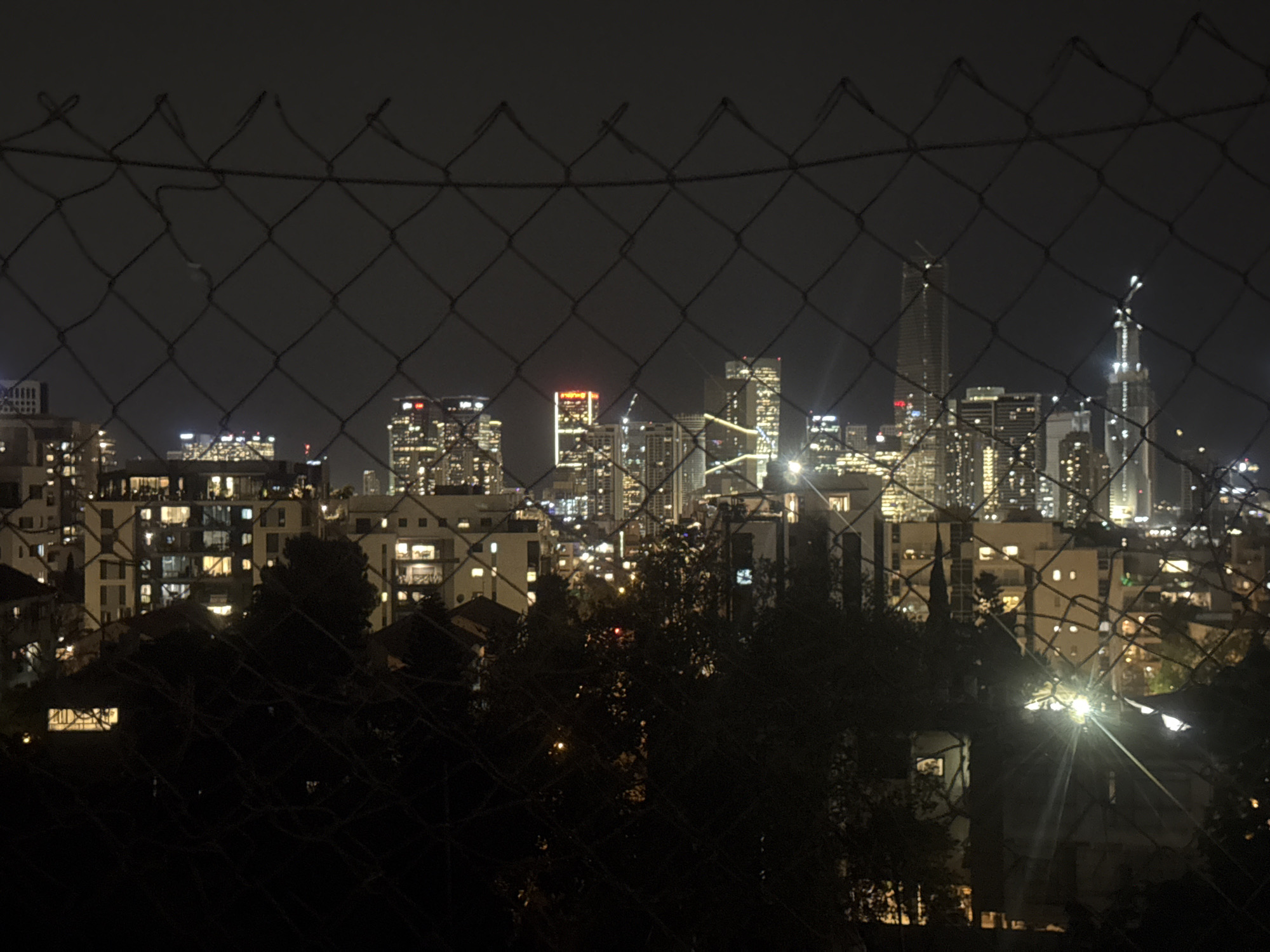 The image shows a nighttime view of a city skyline, likely taken from a residential area in Givatayim, Israel. The cityscape is visible through a chain-link fence, which is the closest element in the frame. Behind the fence, there are residential buildings with lit windows. In the background, a cluster of illuminated skyscrapers can be seen against the dark sky. The buildings appear to be in a downtown or business district of the city. The lighting suggests a mix of residential and commercial activity, with the city lights contributing to the overall ambiance. Some trees are visible in the mid-ground, adding to the depth of the scene.