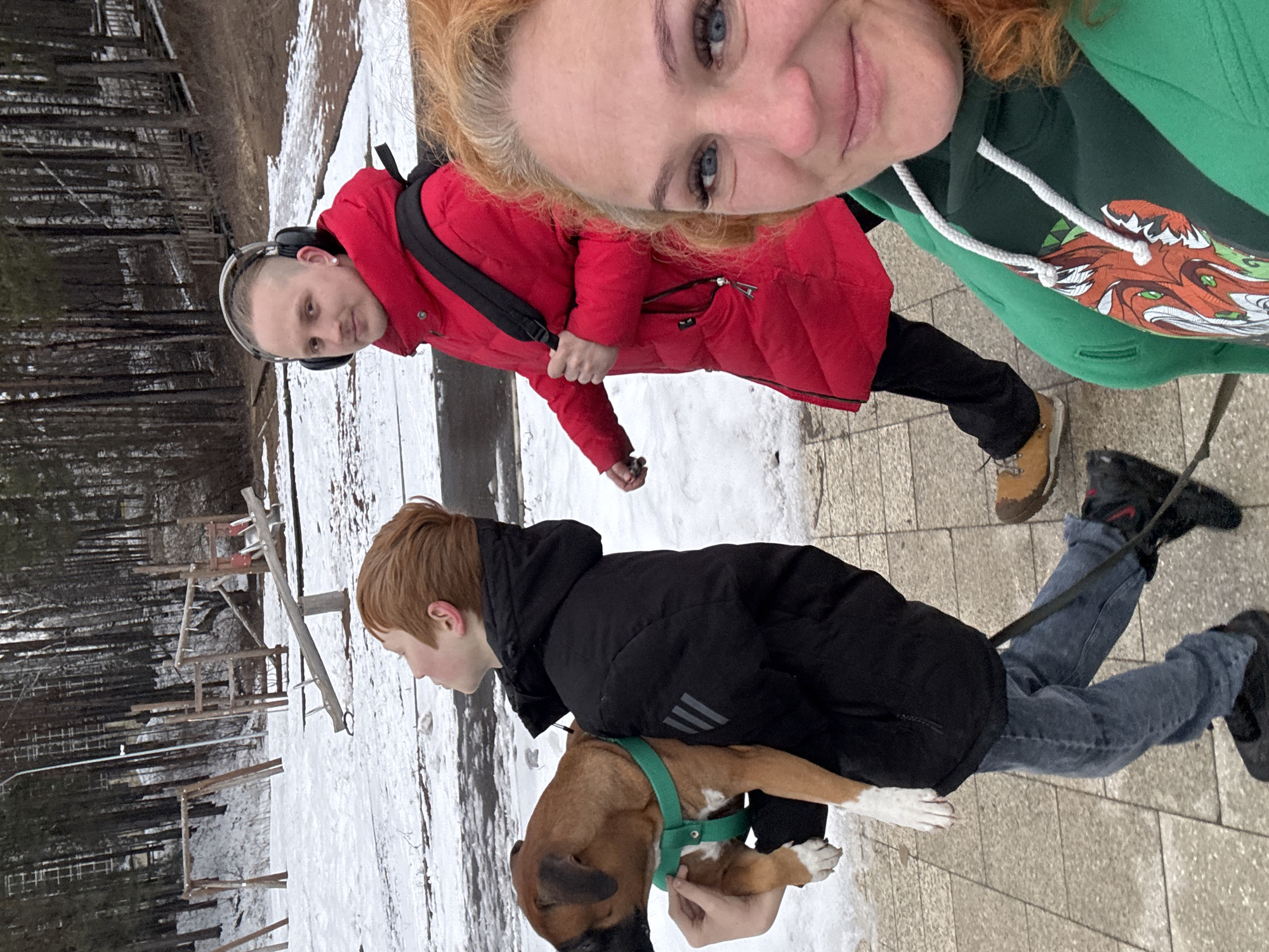 The image depicts three people and a dog in an outdoor setting. A woman with red hair is visible in the upper right corner, wearing a green hoodie. Next to her, a man in a red jacket with headphones around his neck is visible. Below them is a younger person wearing a black jacket, jeans, and black shoes, holding a brown dog with a green harness. The ground is partially covered with snow. The background shows bare trees, possibly a forest or park area, and some playground equipment. The location is Chaikovsky, Russia.
