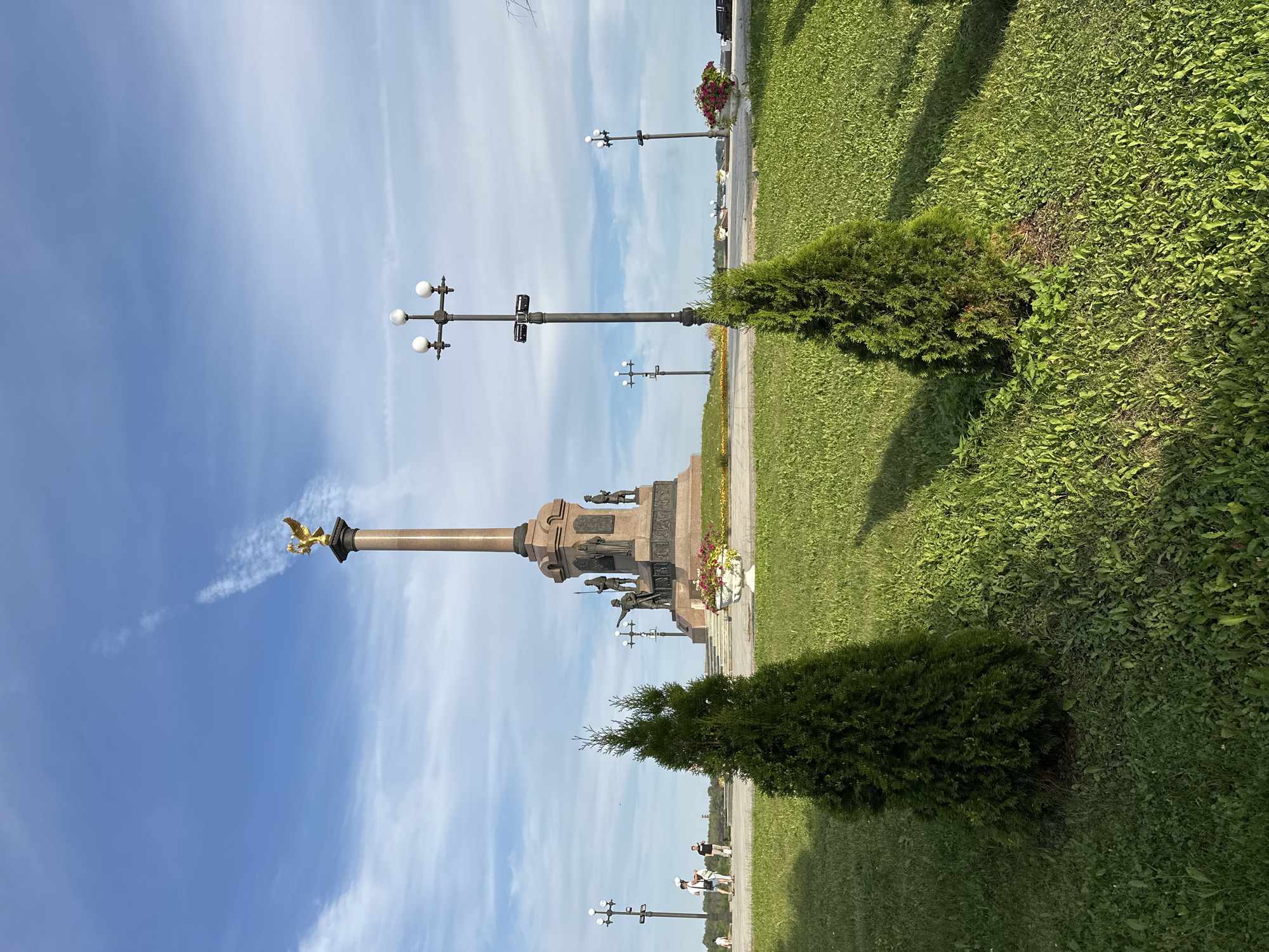 This is a photograph taken in Rusanovo, Russia, featuring a memorial monument topped with a golden eagle. The monument is situated on a raised platform in the background, with steps leading up to it. Several statues of figures in historical attire are visible around the base of the monument. In the foreground, two small evergreen trees are planted on a well-maintained grassy lawn. Several lampposts are visible along a paved walkway behind the lawn, and a few people are walking in the distance. The sky is blue with scattered white clouds, and the overall scene appears to be an outdoor public space, likely a park or plaza.