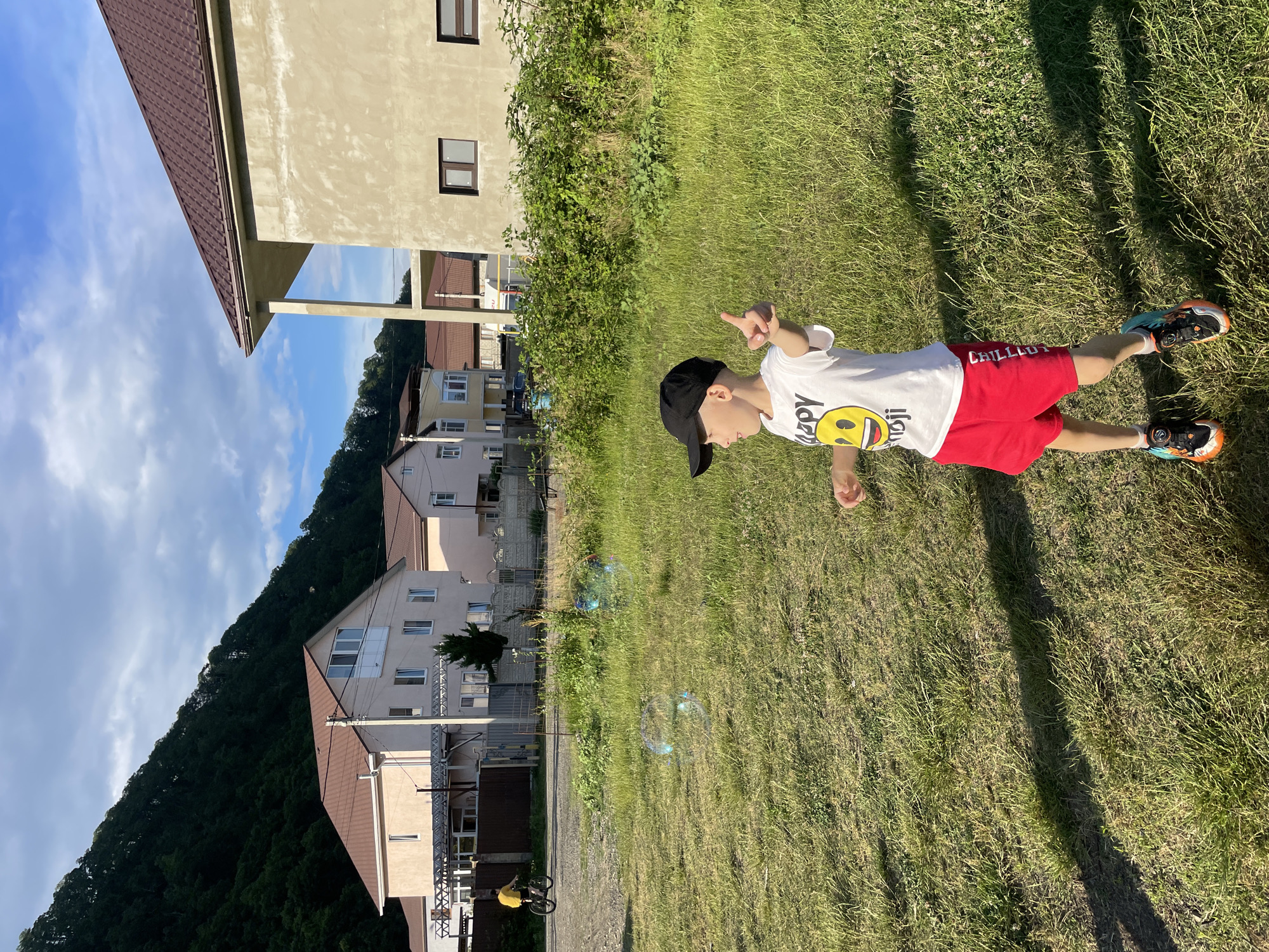 In Baranovka, Russia, on a bright, partly cloudy day, a young boy wearing a black baseball cap, a white t-shirt with a smiley face design, and red shorts stands in a grassy field. He is actively playing, with his right arm extended and pointing forward, and his left arm bent at the elbow, possibly in the process of blowing bubbles, as several iridescent bubbles are visible in the air around him. The boy is wearing athletic shoes. Behind him, houses with brown roofs are visible, set against a backdrop of a densely forested mountainside under a blue sky with scattered white clouds. In the middle ground, a person on a bicycle is seen riding on a gravel path. The overall scene depicts a casual outdoor activity in a residential area near natural scenery.