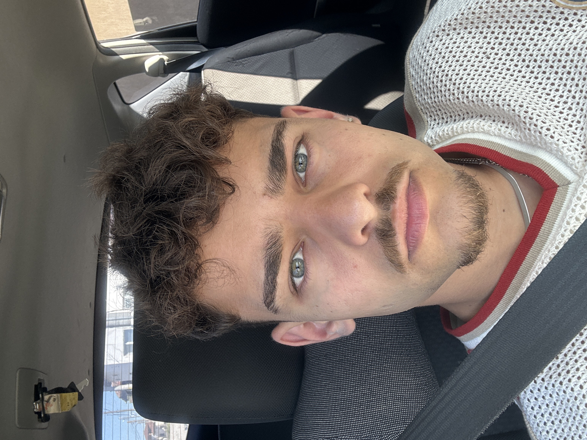 A young adult male is shown in a close-up selfie within a vehicle. The man has light skin, curly brown hair, green eyes, and light facial hair. He is wearing a white, open-weave sweater with red and cream trim around the neck and a silver chain. A black seatbelt crosses his chest. He is sitting in a black car seat with a gray headrest. The car's interior is visible, with the window and door frame in the background. Outside the window, indistinct buildings can be seen. The location is unknown.