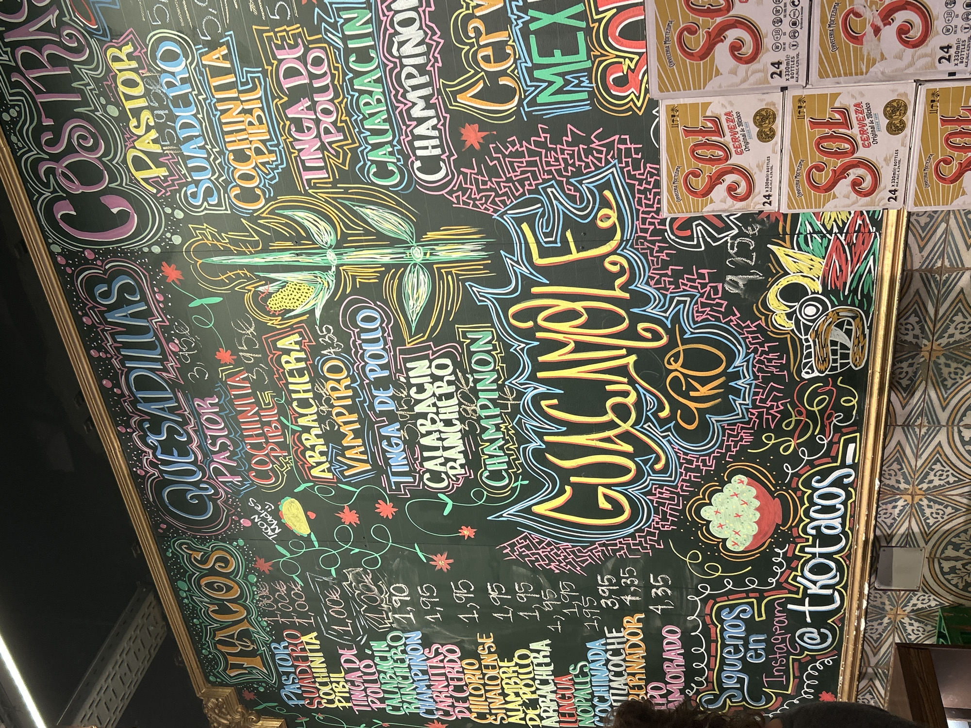 This image shows a close-up, eye-level view of a large chalkboard menu in what appears to be a Mexican restaurant. The chalkboard is decorated with colorful, hand-drawn lettering and illustrations of food items and decorative elements. The menu items listed include various types of tacos, quesadillas, and other dishes with Spanish names, along with prices. In the lower right portion of the image, several cardboard boxes of "Sol Cerveza Original de Mexico" are stacked, indicating that the establishment also serves beer. The wall behind the chalkboard features a decorative tile pattern. The scene suggests a vibrant and casual dining environment. The presence of the specific beer brand, "Sol Cerveza," and the style of the chalkboard menu are consistent with many informal eateries found in Barcelona, Spain.