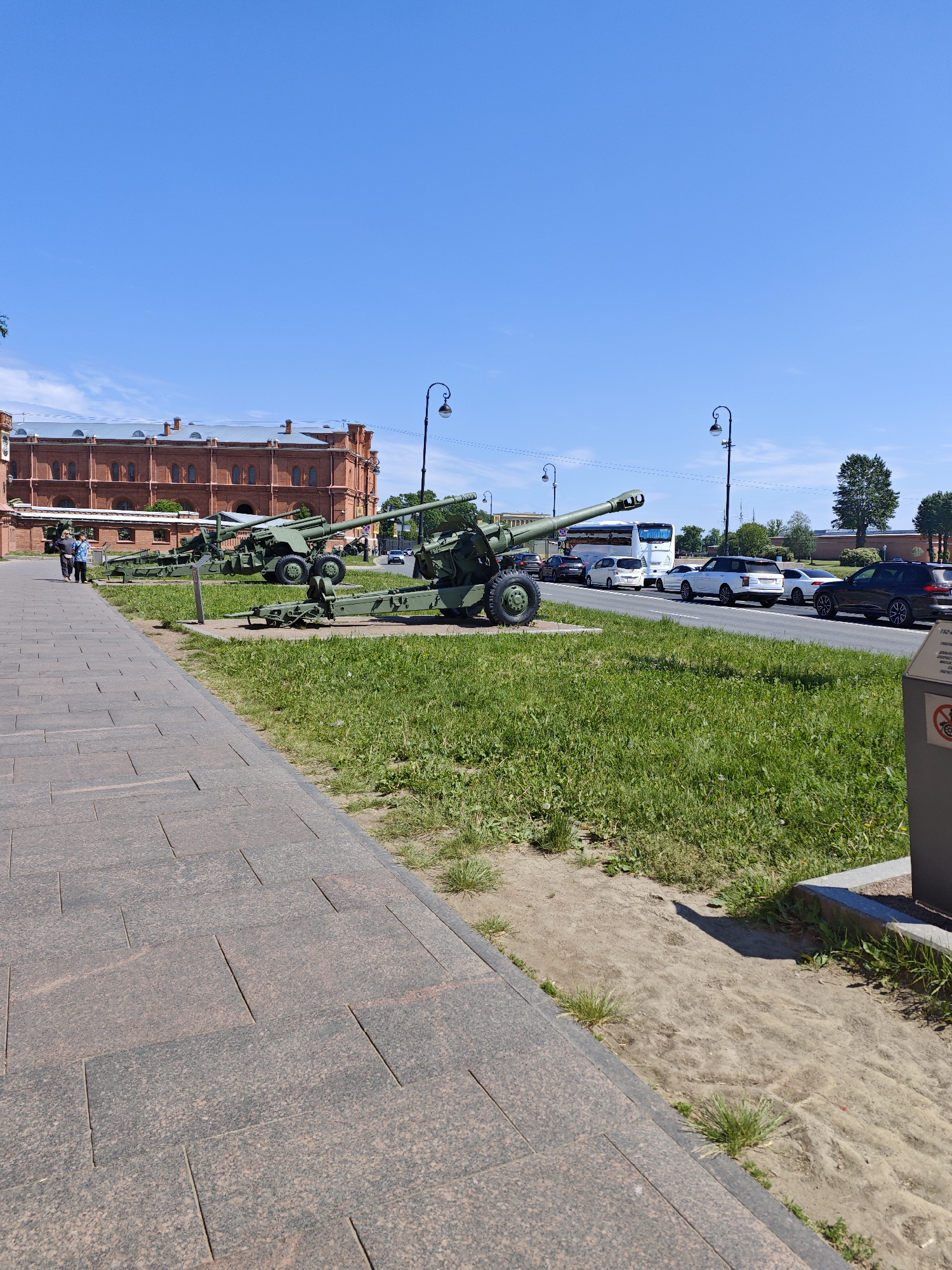 The image depicts an outdoor scene on a sunny day in Saint Petersburg, Russia. In the foreground, a paved pathway made of large, rectangular, dark red and grey granite tiles runs diagonally from the bottom left corner towards the right. Adjacent to the pathway is a strip of green grass with some patches of dry earth and sparse weeds.
Several large, olive-green artillery pieces are positioned on the grass, appearing to be on display. One artillery piece is prominent in the middle ground, facing left, with a large wheel and complex mechanical components visible. Another is positioned behind it and to the left.
In the background, a long, low, red brick building with multiple arched windows and a grey roof stretches across the scene. A blue sky with a few wispy white clouds occupies the upper portion of the image. Across a road to the right of the artillery pieces, there is a line of traffic, including a white bus, several cars, and what appears to be a white SUV. Trees and a section of a wall or building with battlements are also visible beyond the road. Two people are walking on the pathway in the left mid-ground, facing away from the camera. A street lamp with an ornate design is visible on the right side of the image, near the road. A dark grey informational display with some text and symbols is also present in the lower right corner.