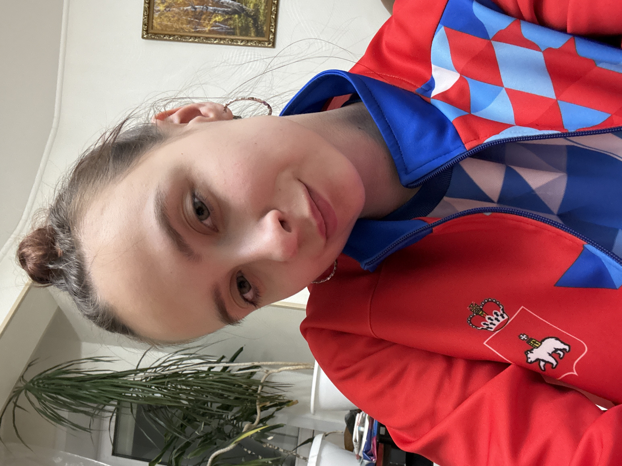 A woman with her hair in a bun is facing the camera. She is wearing a red, white, and blue jacket with a crest featuring a crown and a bear. In the background is a potted plant and a framed landscape painting. The location is Belaya Pashnya, Russia.