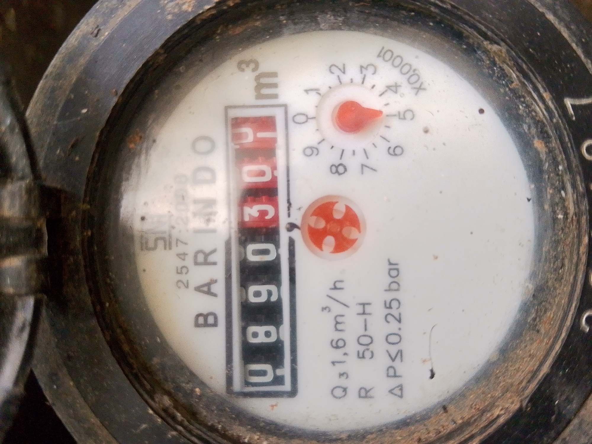 This is a close-up, eye-level shot of a water meter. The meter is round and has a black outer casing that appears to be dirty and worn. The face of the meter is white with black markings and a red digital display. The digital display shows "0.890" with a smaller "3" in a superscript next to it, indicating cubic meters. There are also smaller analog dials with red needles and numbers ranging from 0 to 1000x. Text on the meter includes "BARINDO," "SM," "2547 20-38," "Q₃1,6m³/h," "R 50-H," and "ΔP≤0.25 bar." The background is out of focus and appears to be dark and possibly outdoors. The meter is located in the city of Barabai, Indonesia. There are no people or animals visible in the image.