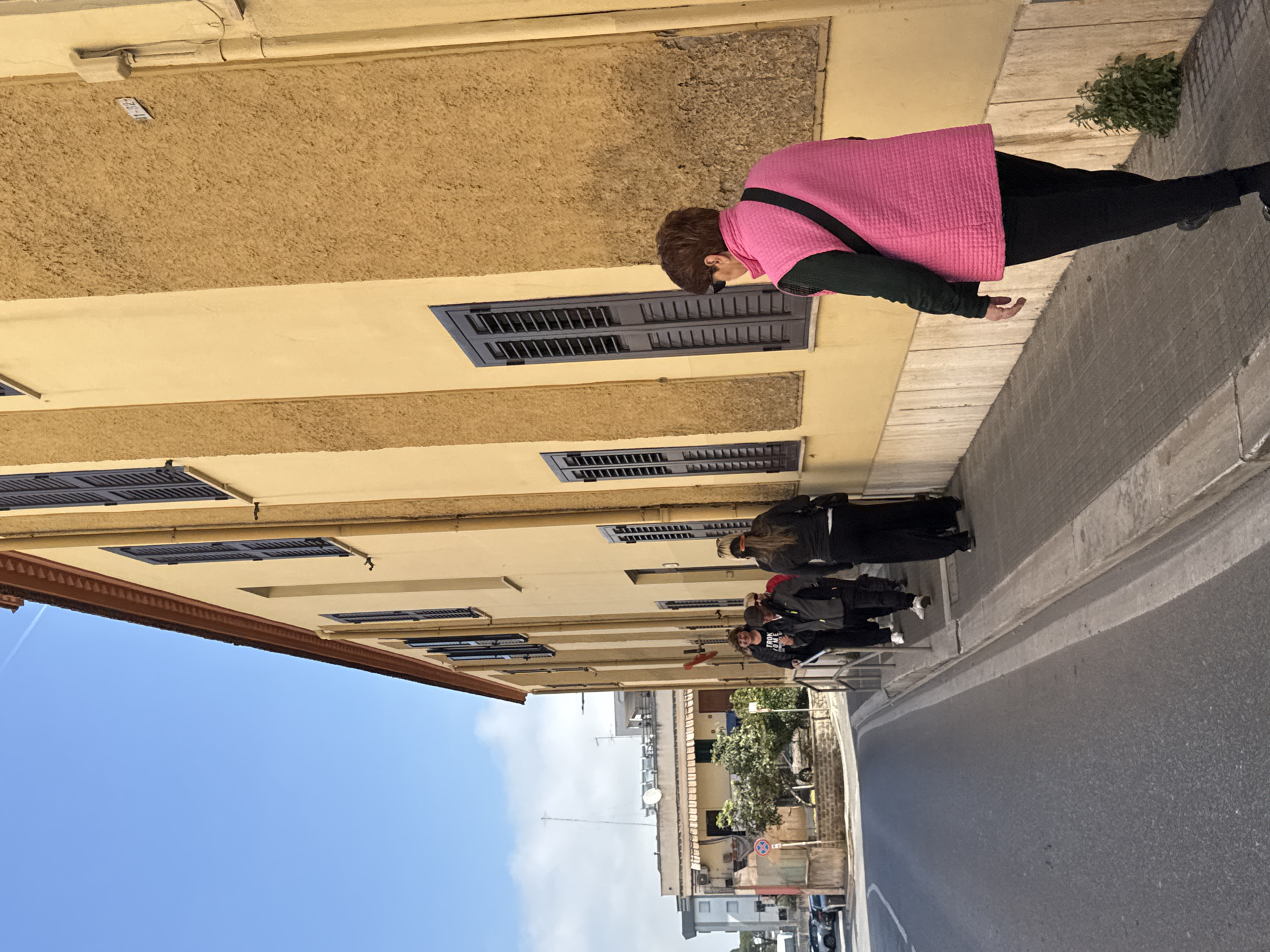 A street scene in Nettuno, Italy, shows a yellow building with dark shuttered windows along a sidewalk. A woman in a pink jacket walks alone near the building's edge. Further down the sidewalk, a group of three individuals walks together. The street runs parallel to the sidewalk, with buildings visible in the distance. A clear blue sky is above.