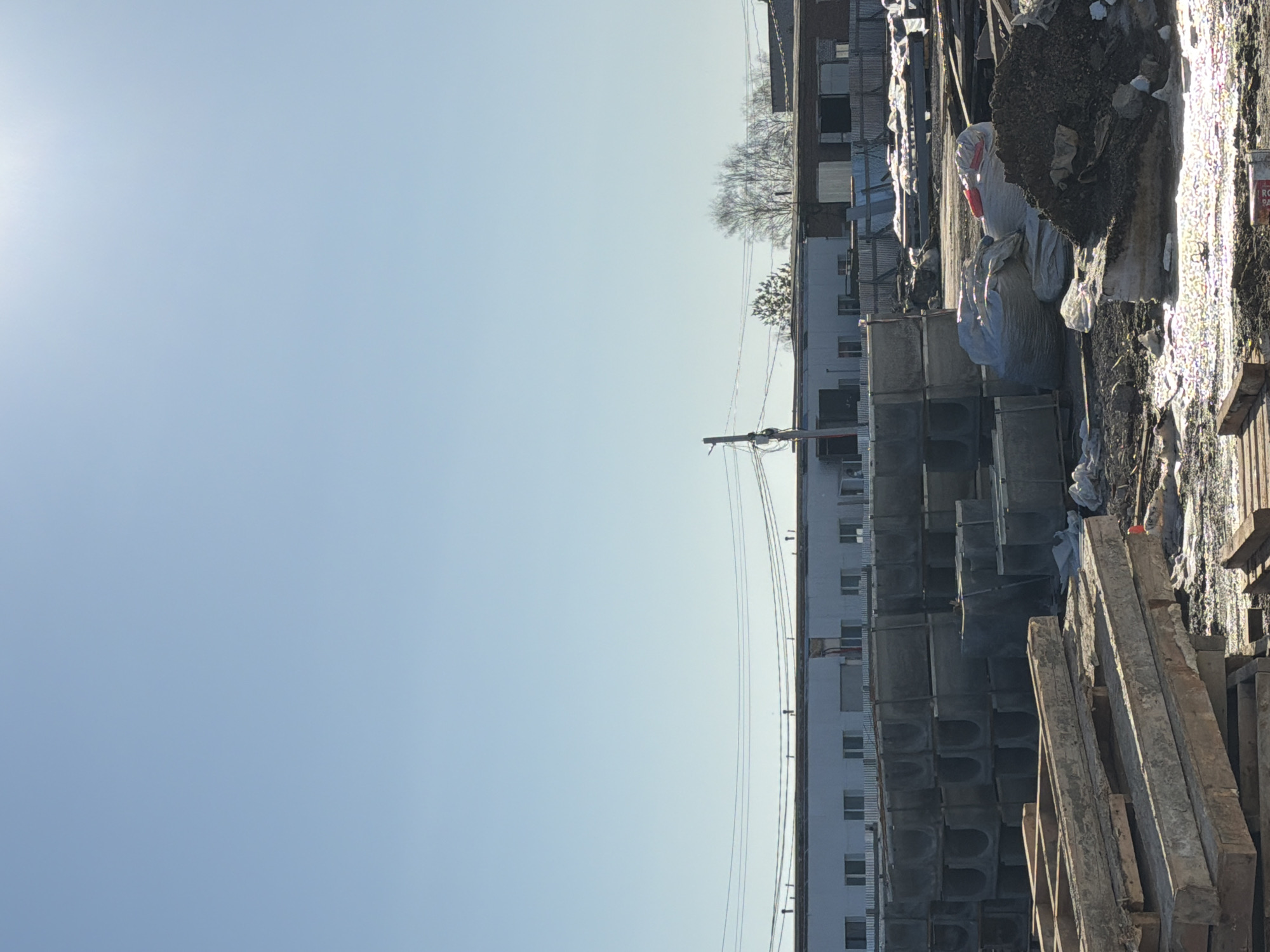 The image depicts an outdoor construction site in Chaikovsky, Russia. A tall residential building is visible, potentially under construction or renovation, with multiple window openings. Construction materials like concrete pipes and wooden planks are stacked in the foreground. Debris and filled bags are present, suggesting ongoing work. Power lines run across the scene. The sky is clear and bright, indicating daylight.