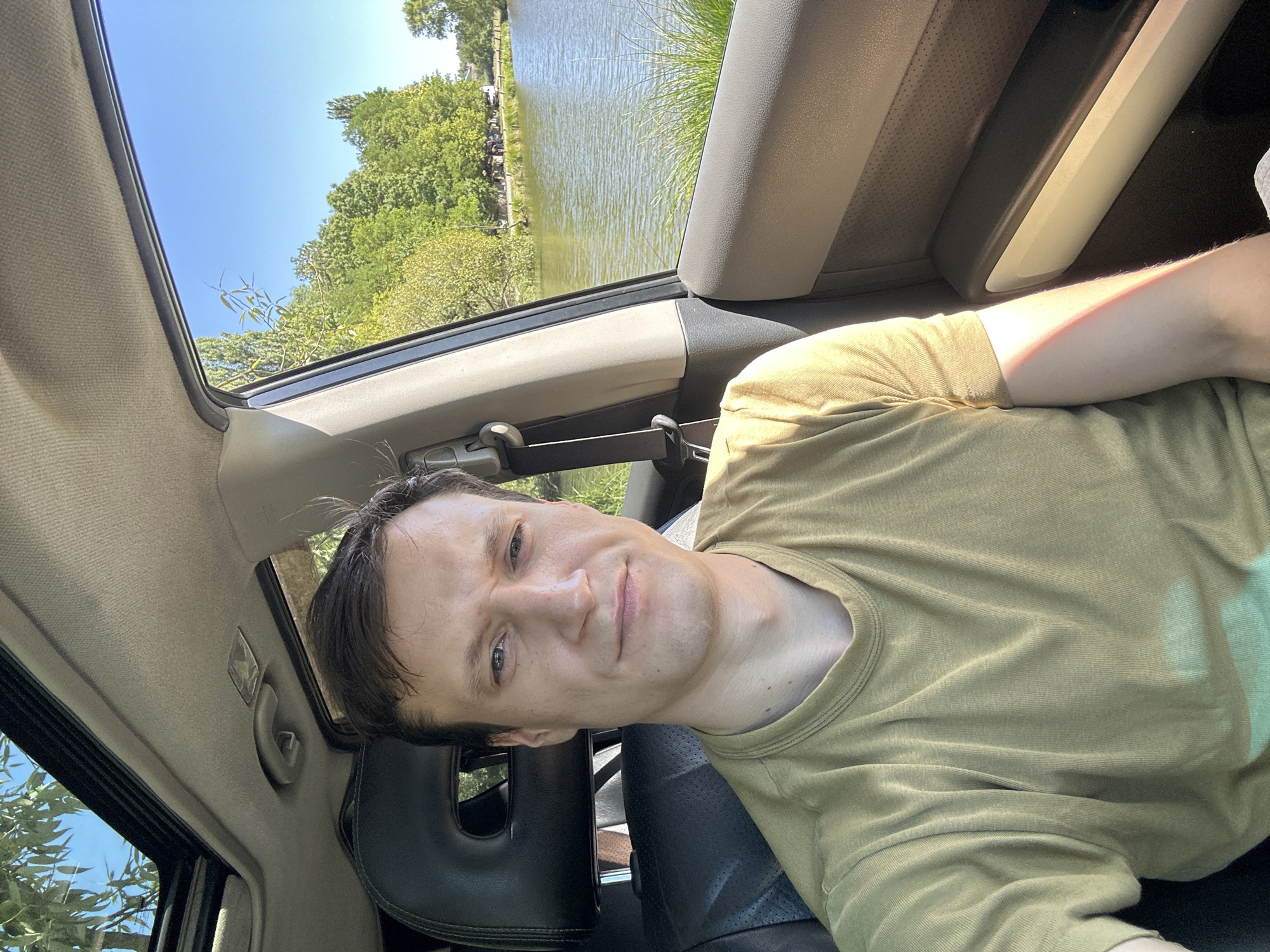 A young man with brown hair and fair skin is sitting in the driver's seat of a car, looking towards the camera with a slight smirk. He is wearing a light olive green t-shirt. Through the open sunroof and window, a sunny day is visible, with a clear blue sky and lush green trees lining a body of water. Several people can be seen in the distance along the shore of the water. The interior of the car is visible, including a black leather seat and a beige headliner. The scene appears to be a park or recreational area, possibly near the village of Makiv, Ukraine.