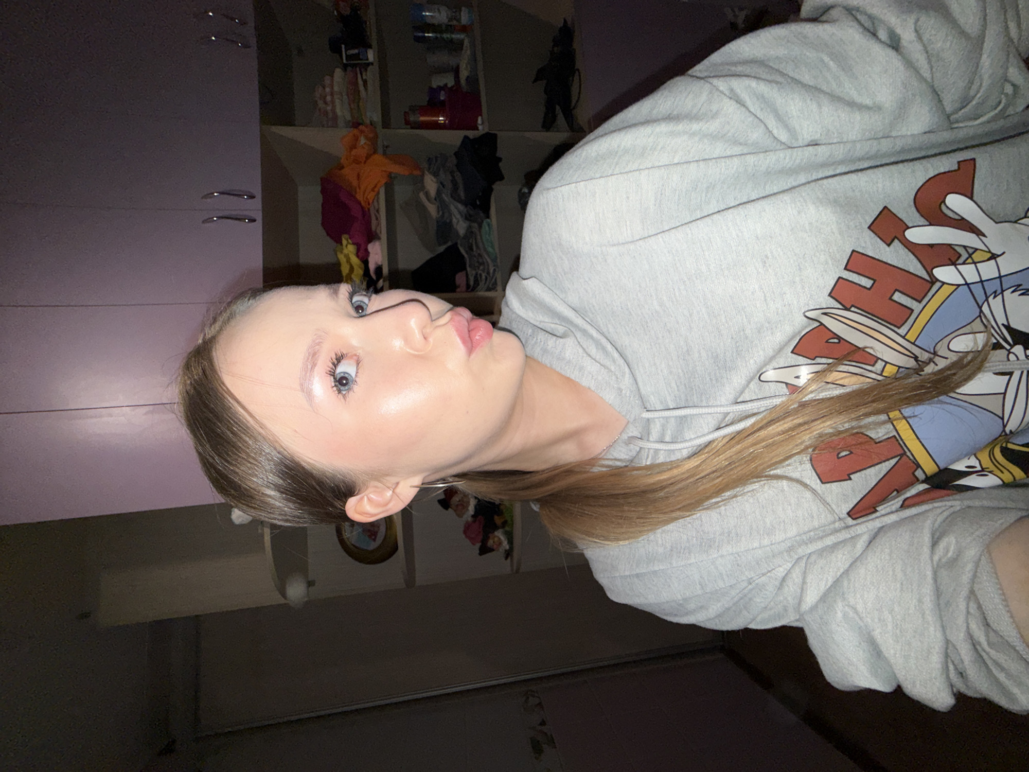 The image shows a young woman indoors, likely in a bedroom or closet. She has blonde hair pulled back into a ponytail and is wearing a grey hoodie with cartoon characters on it. Her face is turned towards the camera, and she is making a kissing face. Her eyes are blue and wide open. The background is somewhat cluttered, with shelves filled with clothes and other items. The lighting appears to be artificial and slightly dim. There is no visible indication that this location is in Zhigalki, Russia.