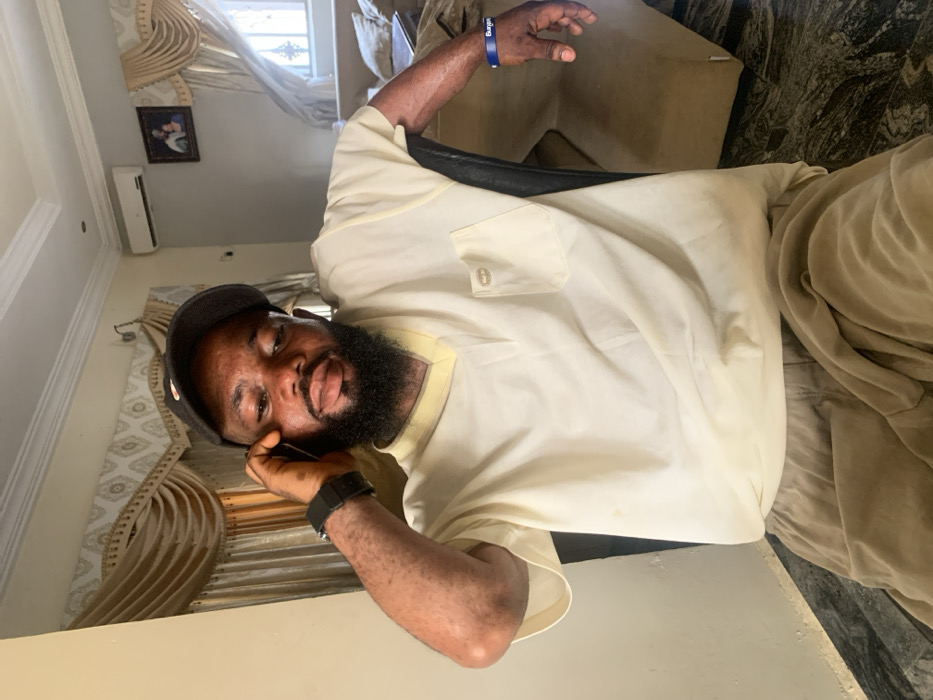 The scene depicts an indoor residential room located in Eika-Adagu New Layout, Nigeria. A single adult male, appearing to be 20-40 years old, with dark skin and a full beard, is seated and facing forward. He wears a black baseball cap, a light cream short-sleeved V-neck shirt with a left chest pocket featuring a small metallic emblem, and light brown trousers. A black digital watch is present on his left wrist, and a blue wristband with white text is on his right wrist. His left hand is raised, holding a dark-colored smartphone to his left ear, indicating he is engaged in a phone call. He looks directly towards the camera. The background consists of light-colored walls, white crown molding, and elaborate gold-toned curtains with valances. The flooring is dark, mottled, and appears to be polished tile. An air conditioning unit is mounted high on the right wall, above a window fitted with security bars. A framed picture hangs on the wall, and a light-colored sofa is partially visible on the right side of the frame. The room is illuminated by ambient light, suggesting a daytime setting.
