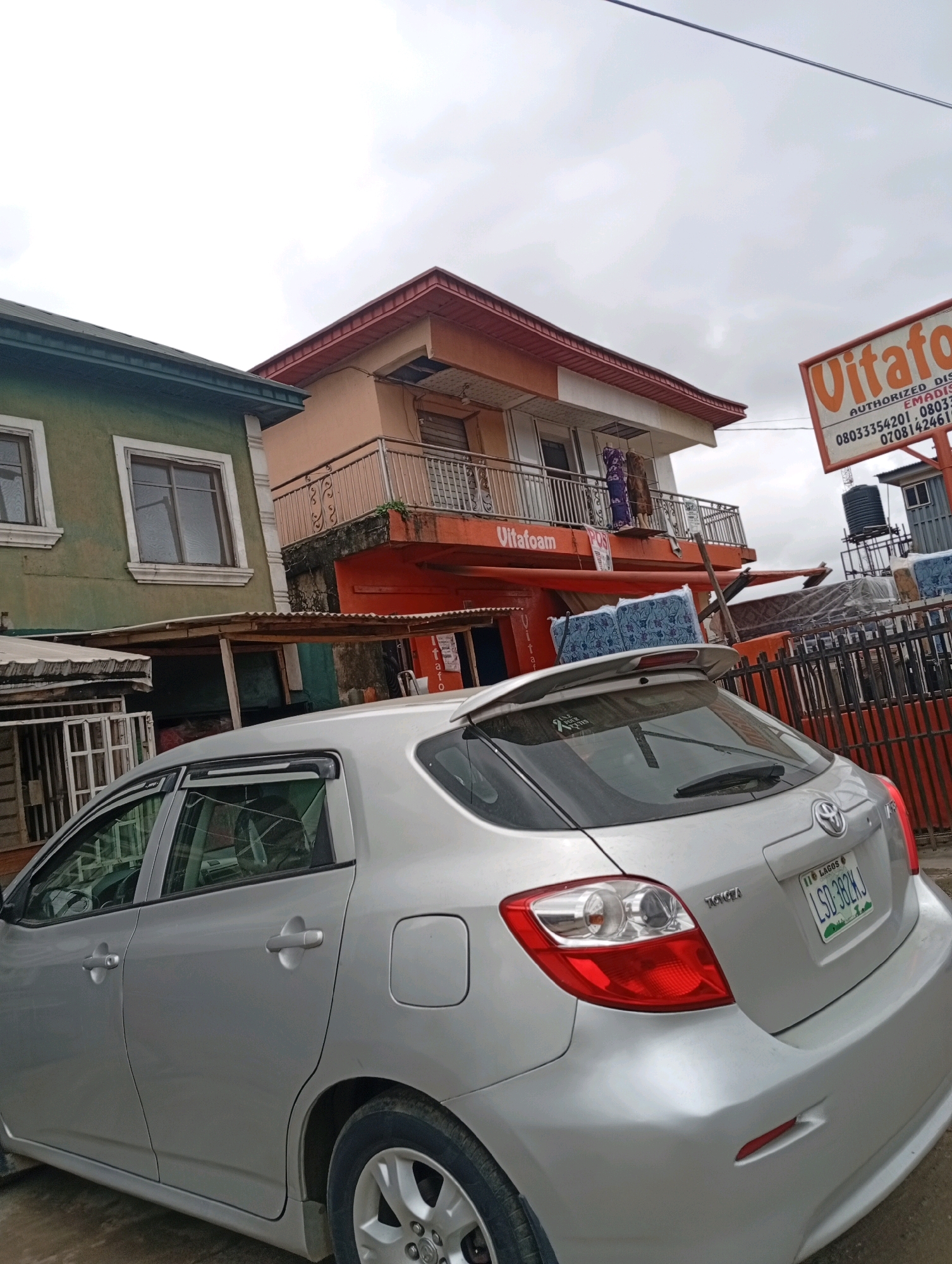 In Akowonjo, Nigeria, a silver Toyota hatchback is parked in front of a multi-story building. The building features an orange facade on its lower levels and a light peach color on its upper story. A balcony with decorative metal railings is visible on the second floor, and laundry hangs from a clothesline on the balcony. A sign for "Vitafoam" is prominently displayed on the building, along with phone numbers. Mattresses are stacked on the ground behind a metal fence. The sky is overcast, and the overall scene suggests a commercial or residential area. The car's license plate reads "LSD 382KJ LAGOS."