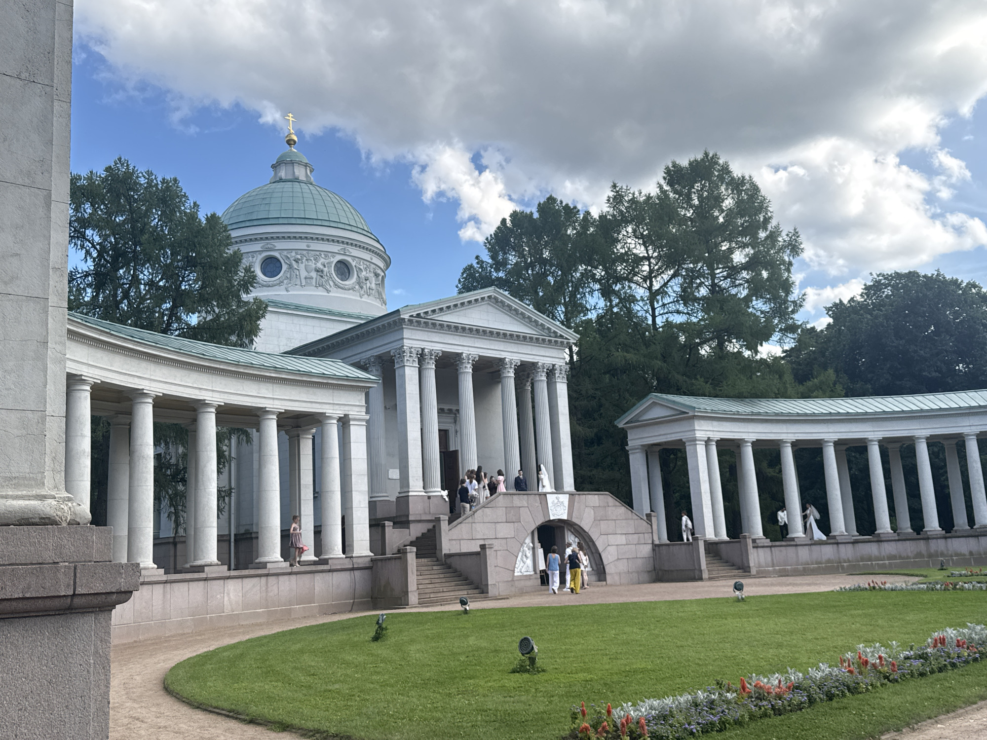 The image shows a neoclassical building with a dome and colonnades, likely a church or a temple, situated in a park. The building is white with a green dome topped by a golden cross. Several people are visible, some walking on a path, others gathered on steps leading to the entrance. There is a well-maintained lawn in the foreground with some flowers. Trees form a backdrop to the building. The sky is partly cloudy with patches of blue. The location is the estate of Arkhangelskoye, in the settlement of Dacha Management Arkhangelskoye, Russia. The scene appears to be a sunny day, and the atmosphere is one of a public gathering or a tourist attraction.