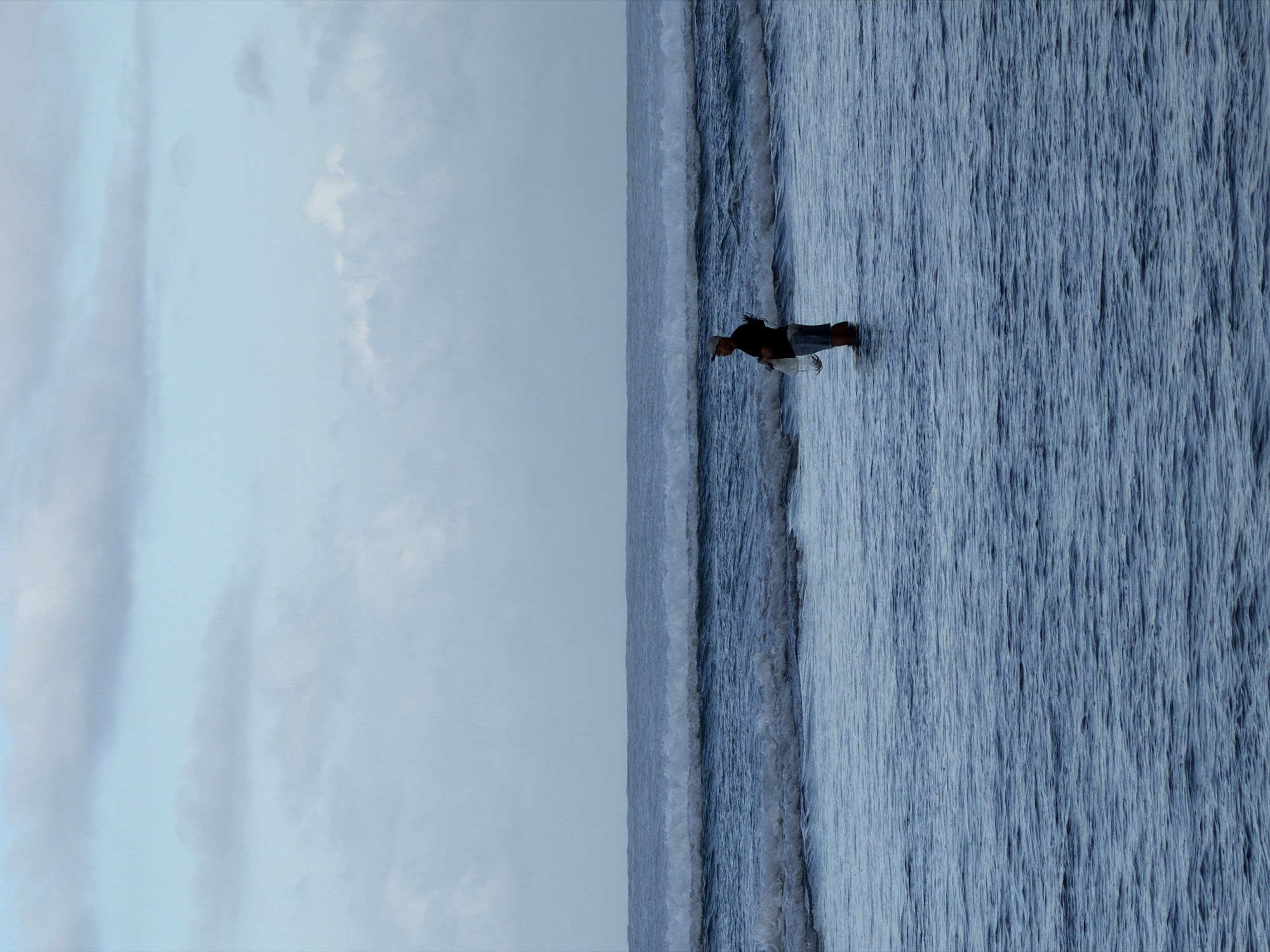 A sideways, eye-level outdoor shot depicts a person in the water on a beach in Kuta Selatan, Indonesia. The person, dressed in dark top, light denim shorts and a light cap, stands in the water with their lower legs immersed, appearing to hold a white object in their right hand. The water, varying shades of blue, exhibits wave patterns, and the sky is light blue with scattered clouds. The horizon is located near the middle, separating the sky and water.