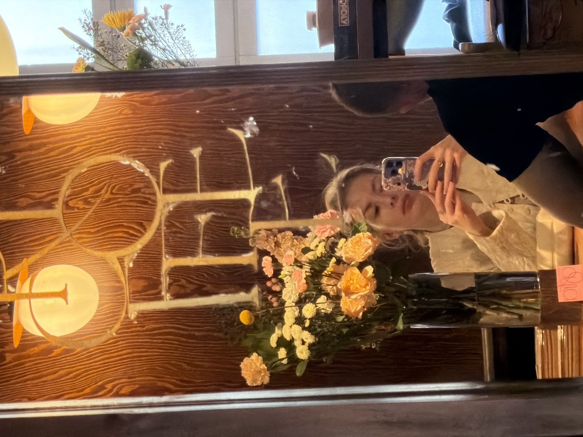 The image is a reflection in a mirror, capturing a woman taking a selfie with her smartphone. She is wearing a beige jacket or shirt. In front of her is a vase of flowers, including yellow and peach-colored blooms. Behind the woman in the reflection is another person, partially visible, wearing a dark-colored shirt. The mirror itself is positioned against a wooden wall with a distinctive circular lighting fixture on the left. Above the mirror, one can see a windowsill with decorative plants and books, one of which has the text "POLY" on its spine. The scene appears to be indoors, possibly a restaurant or cafe in Moscow, Russia.