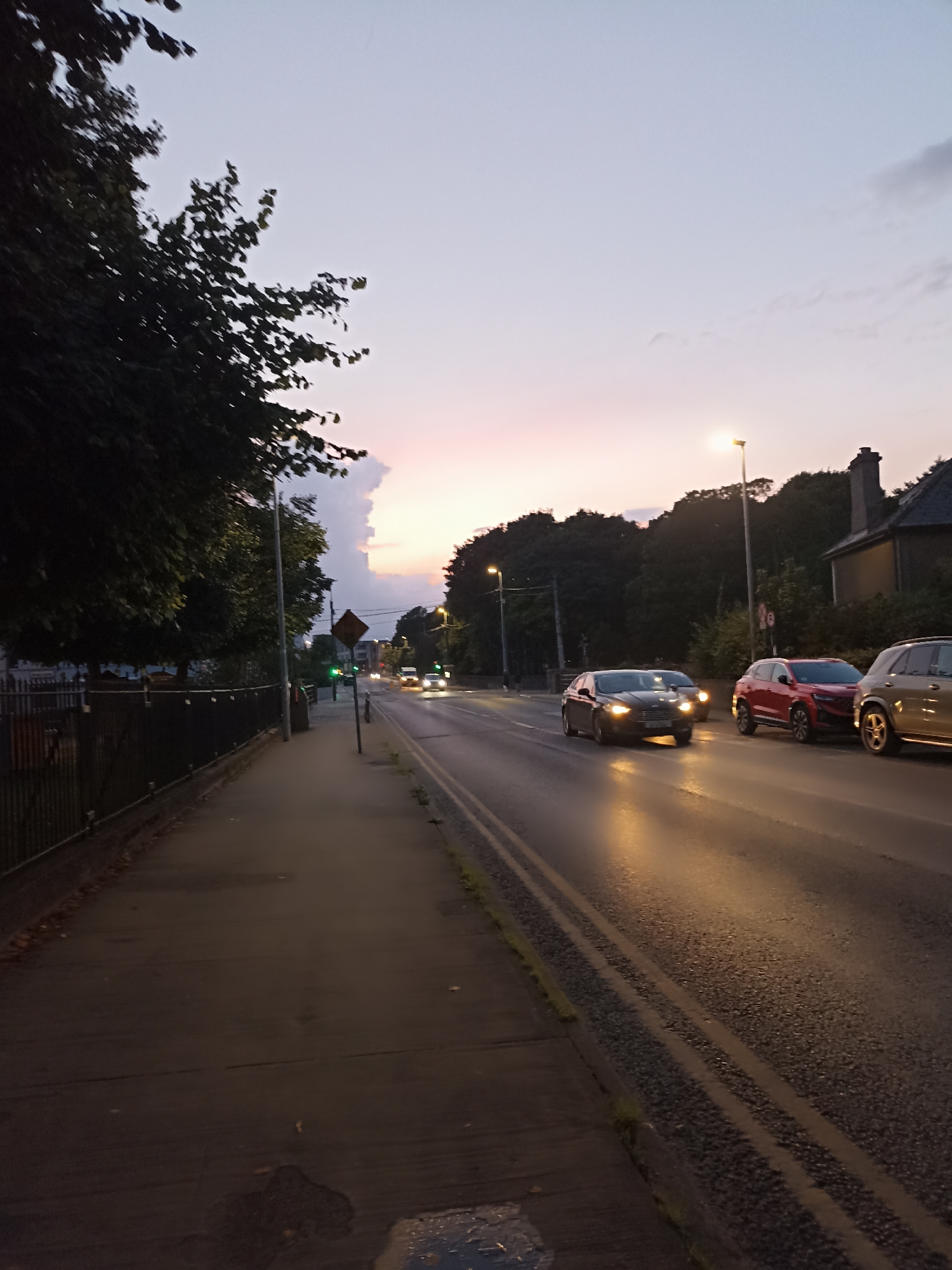 The image depicts a street scene in Galway, Ireland, during dusk. A pedestrian sidewalk runs along the left side of the frame, bordered by a dark metal fence and trees. The sidewalk is paved and appears wet in places. The road is to the right of the sidewalk, with two lanes of traffic visible. Several cars are driving on the road, their headlights illuminated. The sky above is a gradient of pale purple and pink, indicating the setting sun, with some clouds present. Streetlights are visible, casting a warm glow on the wet road. Trees line the far side of the road, and a building with a chimney is partially visible in the background. A traffic light is visible in the distance, showing green. A diamond-shaped traffic sign is also present. The overall impression is of a quiet evening on a city street.