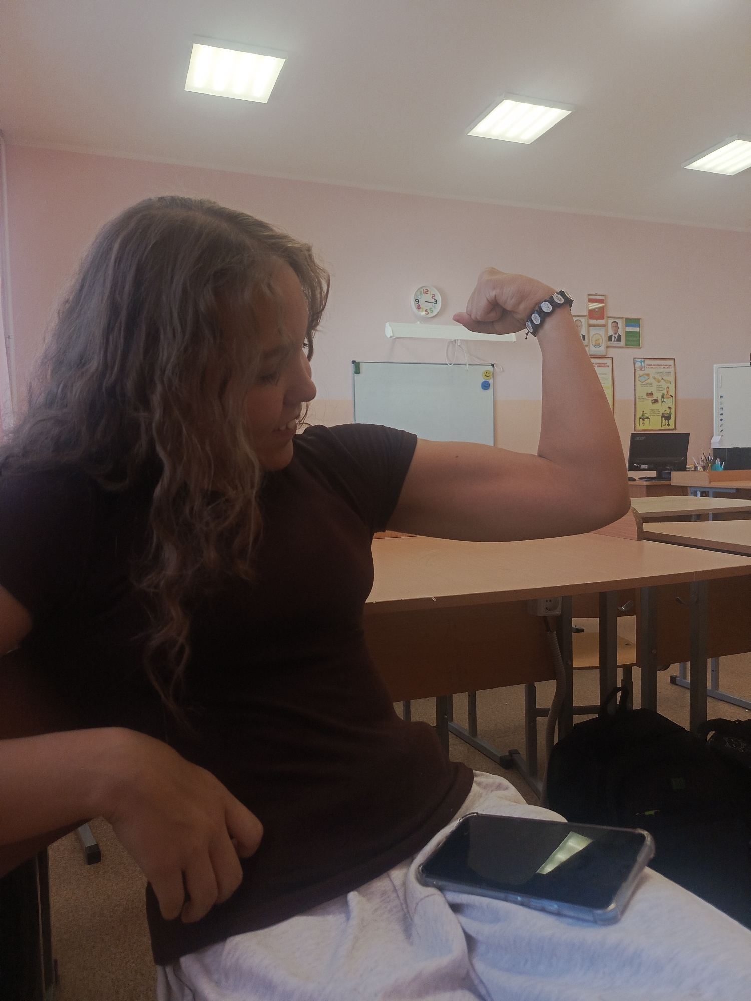 A woman is seated at a desk in what appears to be a classroom. The room has light pink walls and several square ceiling lights. She is wearing a brown t-shirt, and white pants, and a bracelet. A cell phone is on her lap. She is flexing her right arm, exhibiting muscle definition. In the background, there are desks, a whiteboard, a clock on the wall, and some posters. There is also a computer monitor visible on a desk in the background. The location is Unknown, Unknown.