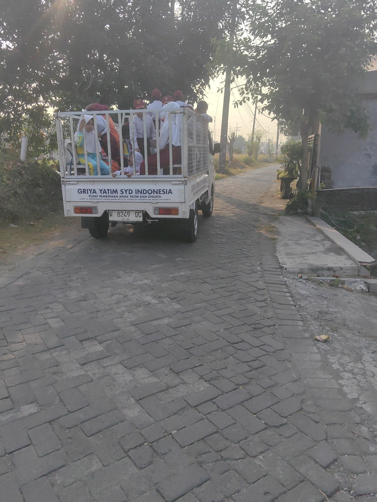 The image shows a white utility truck with a cage in the back, filled with approximately ten children and young adults. The individuals are wearing school uniforms, including red caps and white shirts with some wearing headscarves. The truck has "GRIYA YATIM SYD INDONESIA PUSAT PEMBINAAN ANAK YATIM DAN DHUAFA" printed on its rear. Below this text, an address is visible: "ALAMAT: JL. SRIWIJAYAYAKULIS 9, GANTING, KEC. GEDANGAN, KAB. SIDOARJO". The license plate reads "W 8249 QC". The truck is parked on a paved road with a brick-like pattern. Trees and power lines are visible in the background, along with a building on the right side of the image. The lighting suggests it is either early morning or late afternoon. The scene appears to be in Ganting, Gedangan, Sidoarjo, Indonesia.