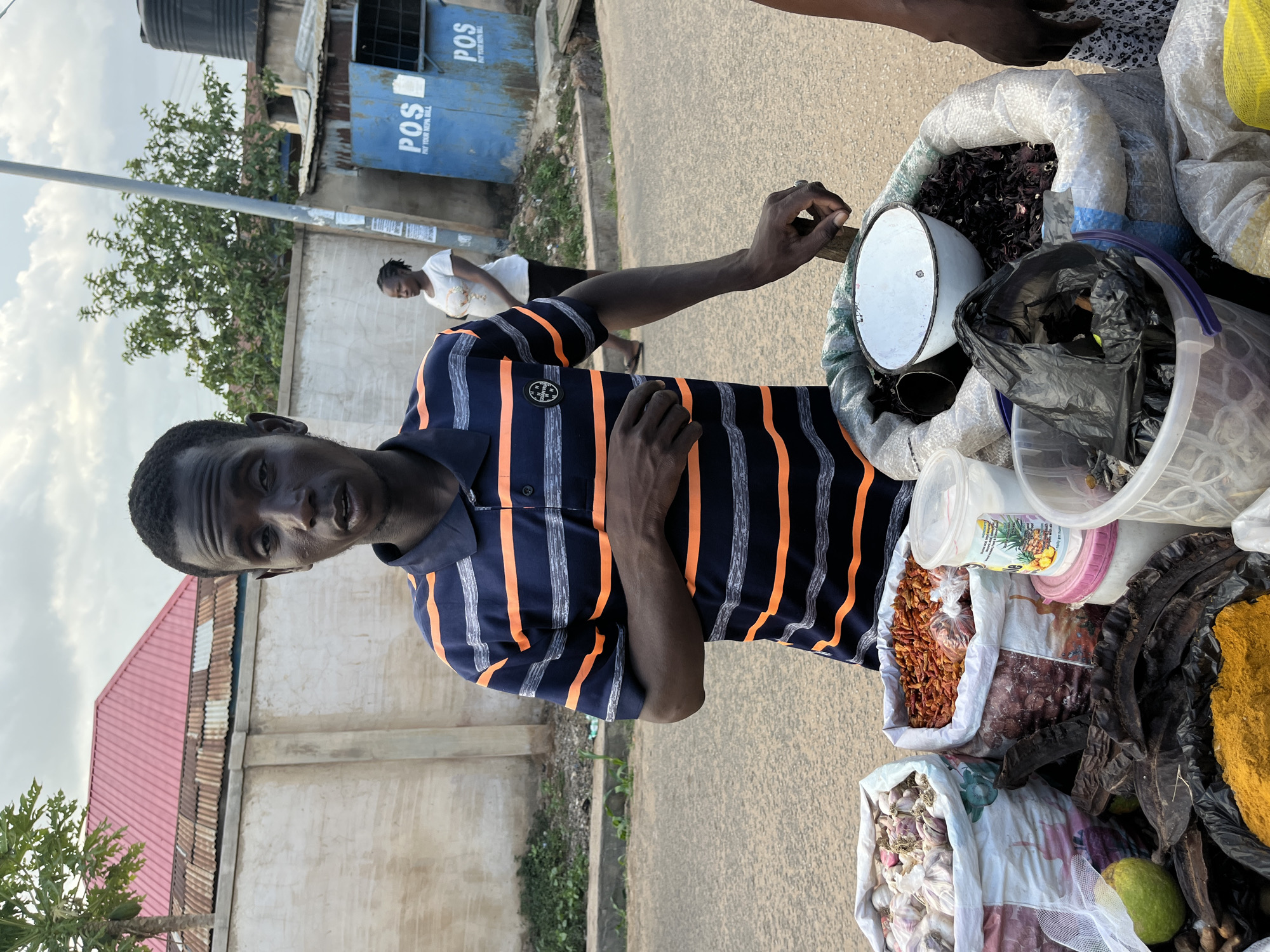 A street vendor is standing next to his goods on a city street. The vendor is a dark-skinned man, wearing a blue and orange striped shirt. The vendor is standing beside a cart filled with goods in sacks, including spices and dried goods. There is a white scale sitting on top of one of the sacks. A woman is walking in the background. The location appears to be in Jos, Nigeria based on the POS business sign in the background.