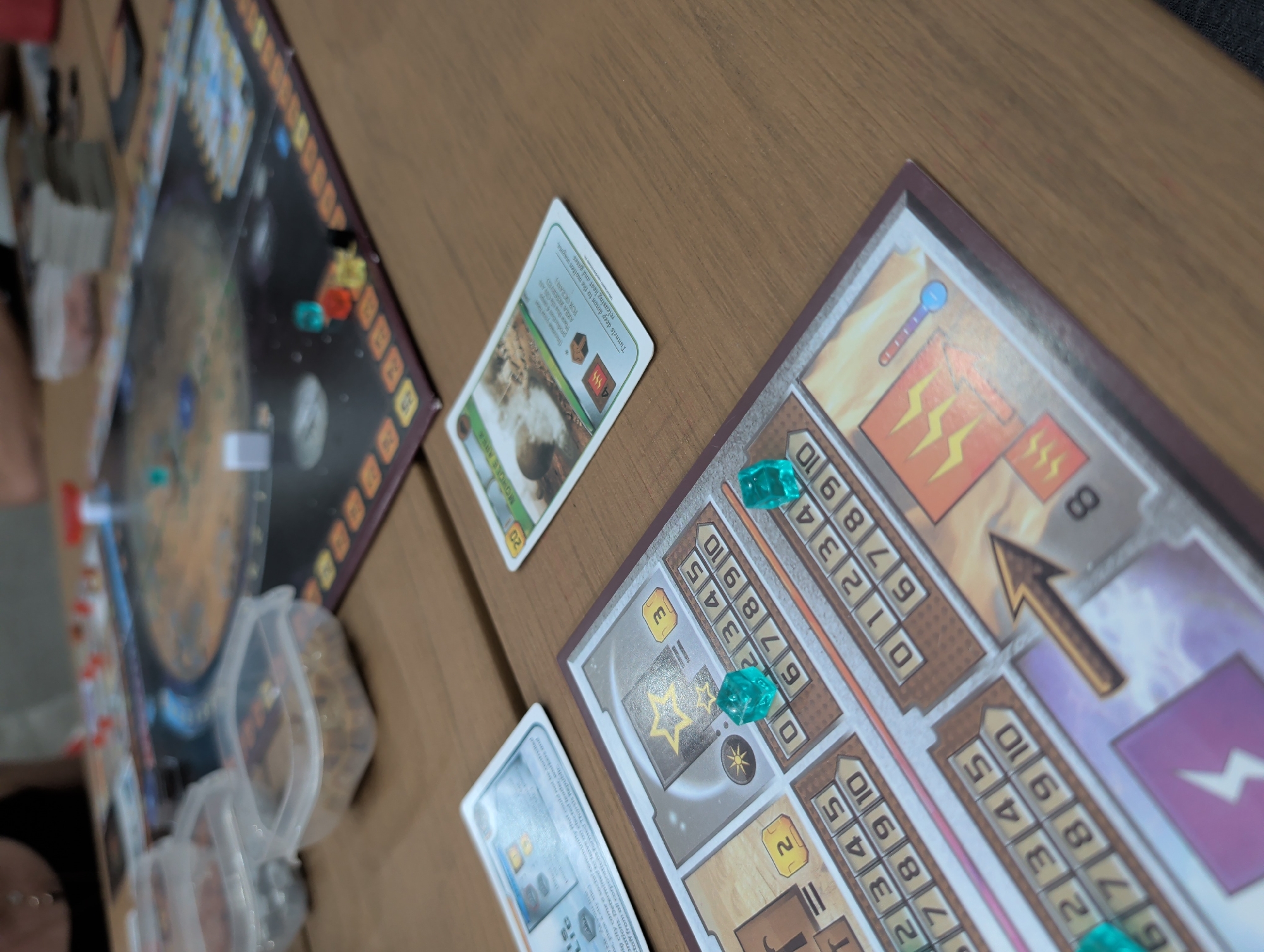 This image shows a tabletop board game in progress on a wooden surface. The components visible include: a circular game board with a Martian landscape and various tokens, cards with text and images, personal player boards with resource tracking, and translucent dice-shaped markers indicating progress along the resource tracks. Specifically, one card shows a location called "MOHOLE AREA". Some cards appear to be discarded or awaiting use. The player boards have different sections for different resources.
