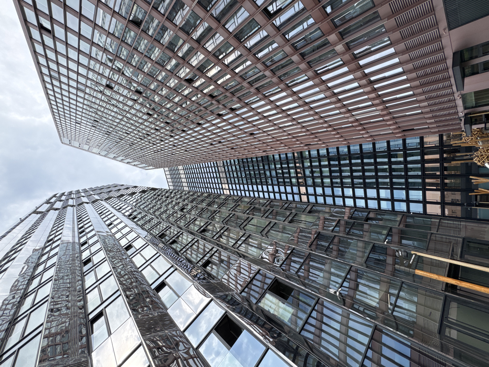 The image shows a low-angle view of modern skyscrapers in Khimki, Russia. The building on the left has a highly reflective, metallic facade with many windows, creating a distorted reflection of the sky and surrounding buildings. The building on the right is clad in a reddish-brown material and also features numerous windows. A third building, with a grid-like pattern of windows, is visible between the two main structures. The sky is overcast. Wooden poles and some abstract metal structures are present in the foreground.