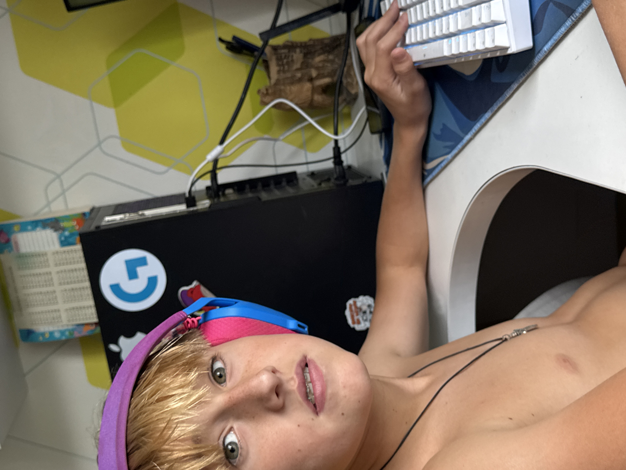 This media file shows a close-up, over-the-shoulder shot of a young person with blonde hair, wearing a purple headband with blue and pink accents. They are lying down, with their head tilted back and their face close to the camera. The person has green eyes, freckles, and braces on their teeth. A thin black cord, possibly from a necklace, is visible across their chest.
The background suggests a gaming setup. A white keyboard with illuminated keys is prominent, placed on a blue and white patterned mousepad. A black electronic device, possibly a computer tower or audio equipment, is visible behind the person's head, with cables connected to it. A circular blue logo with a white "G" shape is on the black device. A piece of textured wood is also present in the background, along with a wall with a geometric yellow and white pattern.
The lighting appears to be indoor, and the overall mood seems casual and relaxed, perhaps during a gaming session. No specific time of day or weather conditions are discernible. No legible text is visible beyond the logo on the device. The context provided indicates the location is Zaporizhzhia, Ukraine.