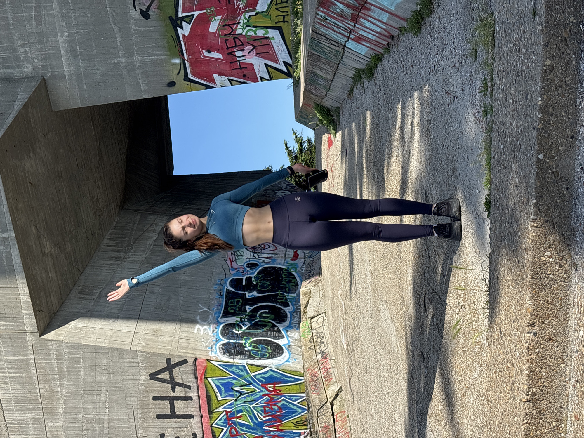 A woman is standing in a passageway under a concrete structure, possibly a bridge or overpass. She is wearing a long-sleeved blue crop top and dark leggings, with black sneakers. Her left arm is extended to the left, and her right arm is bent, holding a dark object, possibly a phone. The walls of the passageway are covered in graffiti. The ground is made of small stones and is partially covered in shadows. The sky is visible through the opening of the passageway. The location is Varna, Bulgaria.