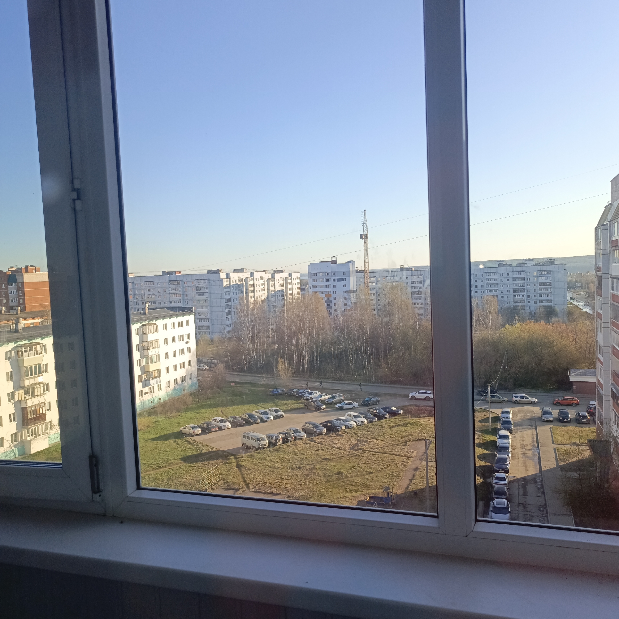 The image presents a view from an interior window, looking out onto an urban or suburban landscape under a clear, bright sky. The window frame, made of white plastic, occupies the immediate foreground, with visible mullions dividing the view and a sill at the bottom.
Beyond the window, a sprawling residential area unfolds. Numerous multi-story apartment buildings, primarily light-colored (white, gray, and light blue), dominate the scene, suggesting a densely populated environment. Their architectural style points to post-Soviet-era construction, characterized by repetitive block designs and numerous balconies. One building on the far right has distinctive reddish-orange sections.
In the midground, between the buildings, are patches of open land. Several unpaved or dirt parking areas are visible, packed with a variety of parked cars, vans, and SUVs. These areas are surrounded by grassy sections, some appearing dry or sparse, and numerous bare deciduous trees, indicating either late autumn or early spring. A paved road or path runs through the scene, with more parked cars lining its edge. A few small, distant human figures can be seen walking on the path, suggesting everyday activity.
A tall construction crane stands prominently in the middle-distance, rising above the bare trees and apartment blocks, hinting at ongoing development or construction in the area. The sky is a clear, brilliant blue, with no clouds visible. The sunlight is strong and direct, casting long shadows across the ground and illuminating the facades of the buildings with a warm glow, suggesting either early morning or late afternoon. No discernible text is visible within the image.