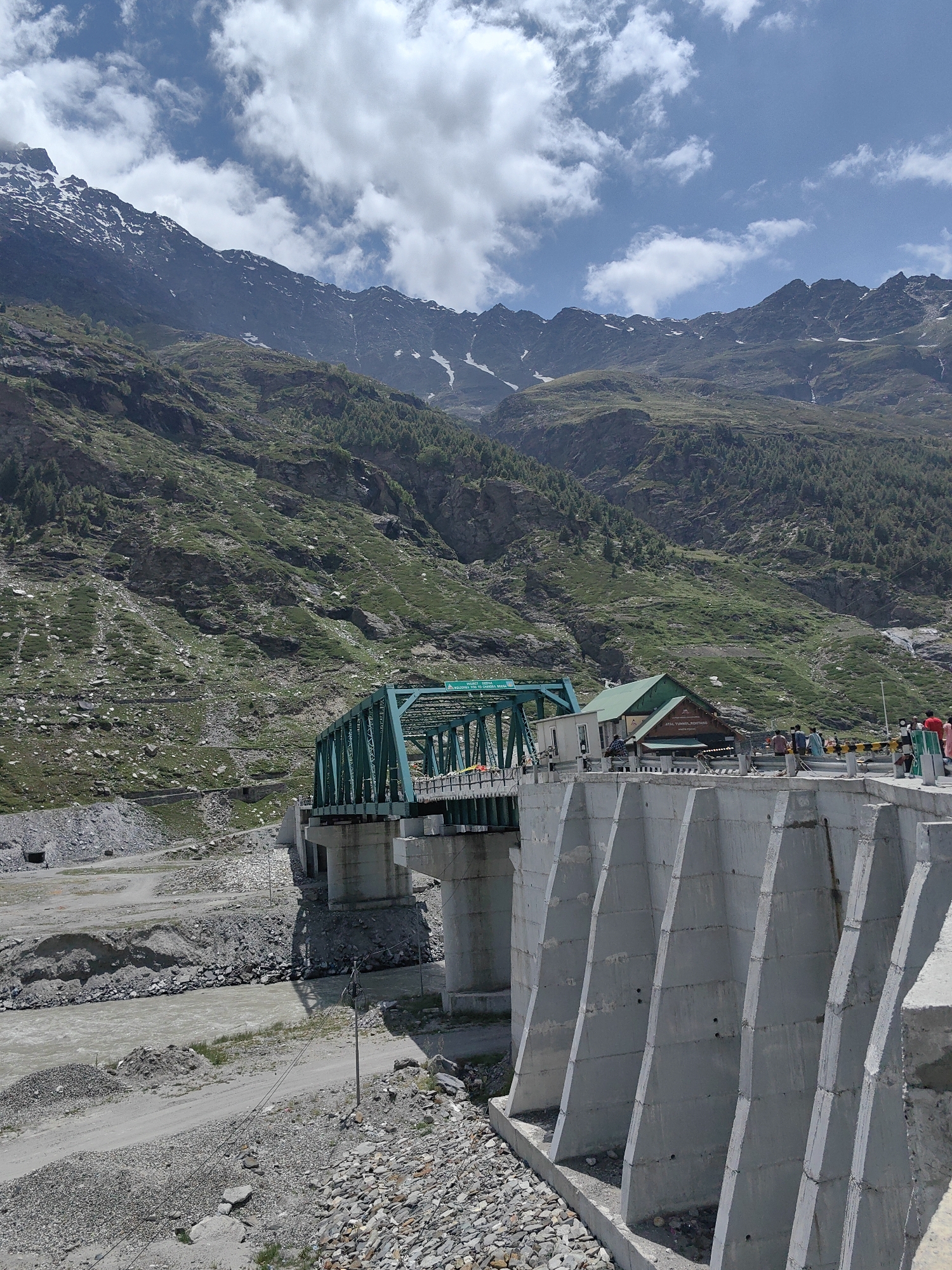 The image depicts a concrete dam structure on the right, with a green truss bridge spanning a river in the background, likely near the city of Burwa, India, given the context. The dam is constructed with large, trapezoidal concrete segments. Beyond the dam, the green truss bridge supports a road and a small building. Several people are visible on the bridge and near the building, appearing to be tourists or locals.
The environment is mountainous, with steep, green slopes covered in vegetation and scattered rocks, rising to jagged peaks in the distance. Some of the higher peaks show patches of snow. The sky is blue with scattered white clouds. A muddy river flows beneath the bridge. The ground in the foreground consists of gravel and rough terrain. The scene suggests a scenic, possibly tourist-oriented location within a mountainous region. The presence of the dam and bridge indicates infrastructure development in the area.