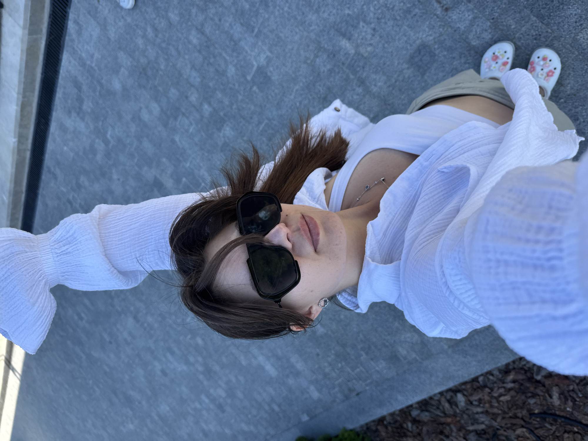 A young woman is captured in a selfie taken from a high angle, looking down at the camera. She is outdoors, as indicated by the bright lighting and the stone-like ground beneath her. The woman has long, dark hair and is wearing large, dark sunglasses, a white tank top, and a white, crinkled blouse with billowy sleeves. She is also wearing khaki shorts and white clogs adorned with pink and red floral patterns. A delicate silver necklace with dark stones is visible around her neck. The setting appears to be a paved area, possibly a patio or walkway, with a dark grate running along the left side of the frame and some mulch or wood chips visible in the lower right corner. The lighting suggests it is daytime, likely sunny. The woman's expression is neutral, and the overall mood of the image is casual and relaxed, possibly capturing a moment of leisure or a planned outfit shot. There is no visible text in the image.