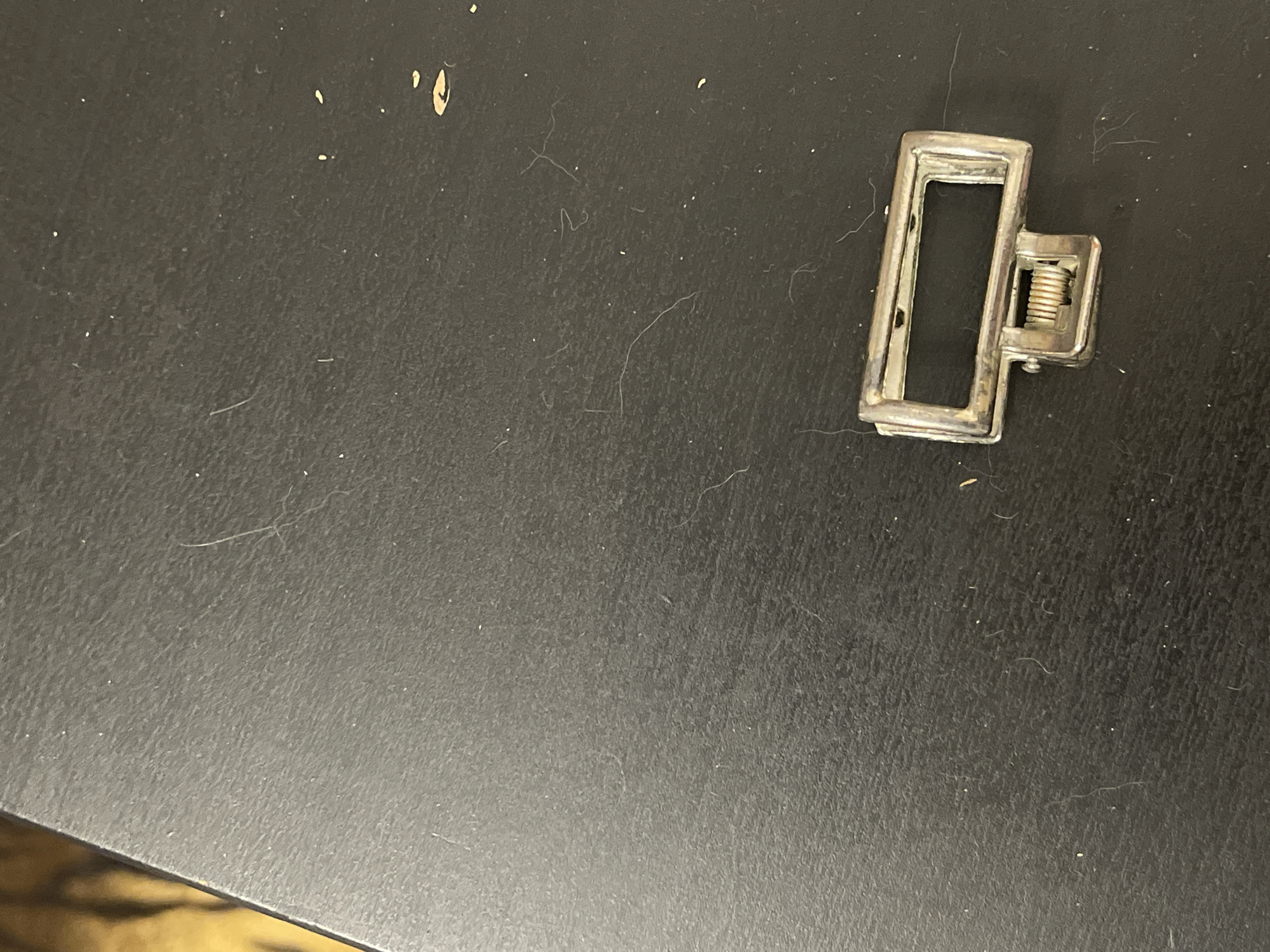 An overhead shot shows a silver colored metal clasp with a spring mechanism resting on a textured black surface. The surface is speckled with white debris and several long, thin strands of white hair or fiber. The clasp is rectangular with a smaller, hinged rectangular piece attached to the bottom center of the longer rectangle. The hinged piece has a coiled spring visible within its structure. The lighting is dim and casts faint shadows.