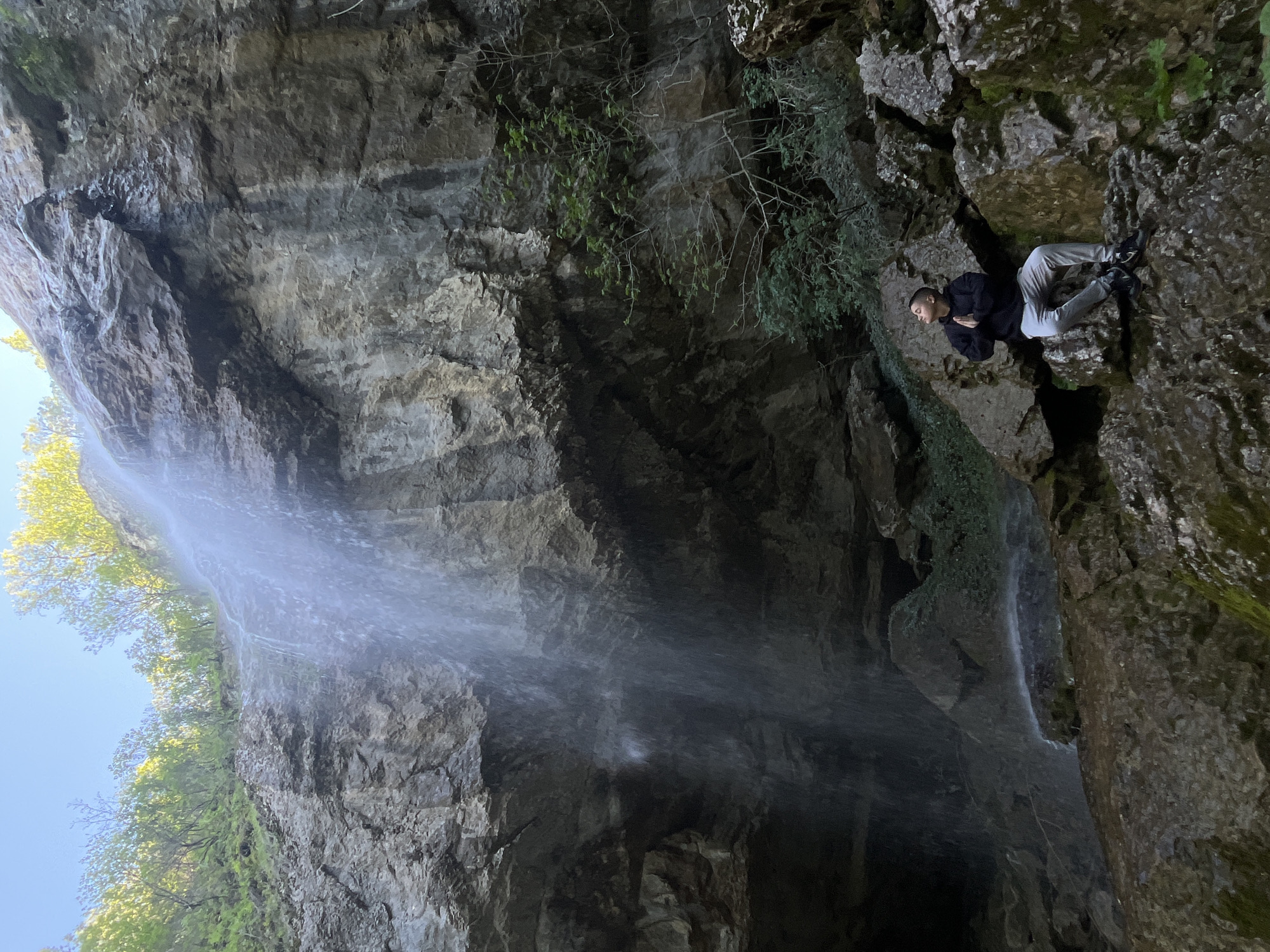 The image shows a person sitting on a rock face next to a waterfall. The location is identified as being in Veselinovo, Bulgaria. The person is wearing a dark jacket and light-colored pants. The waterfall appears to be partially obscuring a rock formation. The surrounding environment includes rocks, moss, and vegetation.