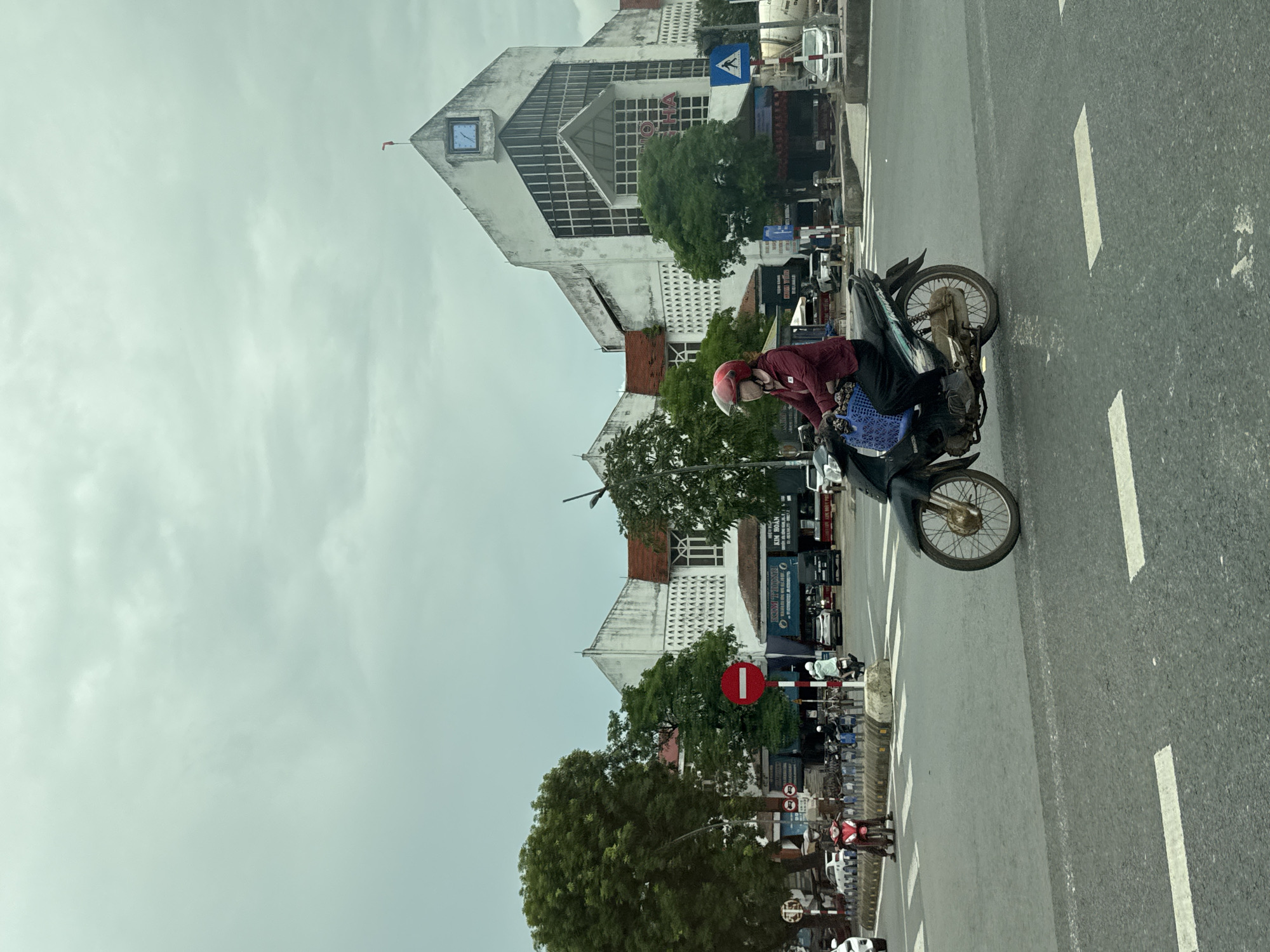 A medium-shot, eye-level, outdoor, daytime, vertical photograph captures a street scene in Nam Đông Hà Ward, Vietnam. The foreground features an asphalt road with white lane markings. A person wearing a red helmet and a maroon jacket rides a dark green motorcycle across the road, heading towards the right. The motorcycle has a blue plastic basket attached to the front. In the middle ground, a pedestrian crossing is visible with a red circular traffic sign indicating prohibition. Several other motorcycles and cars are present on the road and sidewalk. Behind the road, a large, multi-story white building with a distinctive architectural style stands out. The building has a triangular roof with a clock face and a flag on top. There are trees and other structures flanking the main building, suggesting a commercial or public area. The sky is overcast with visible clouds, indicating a dull weather condition.
