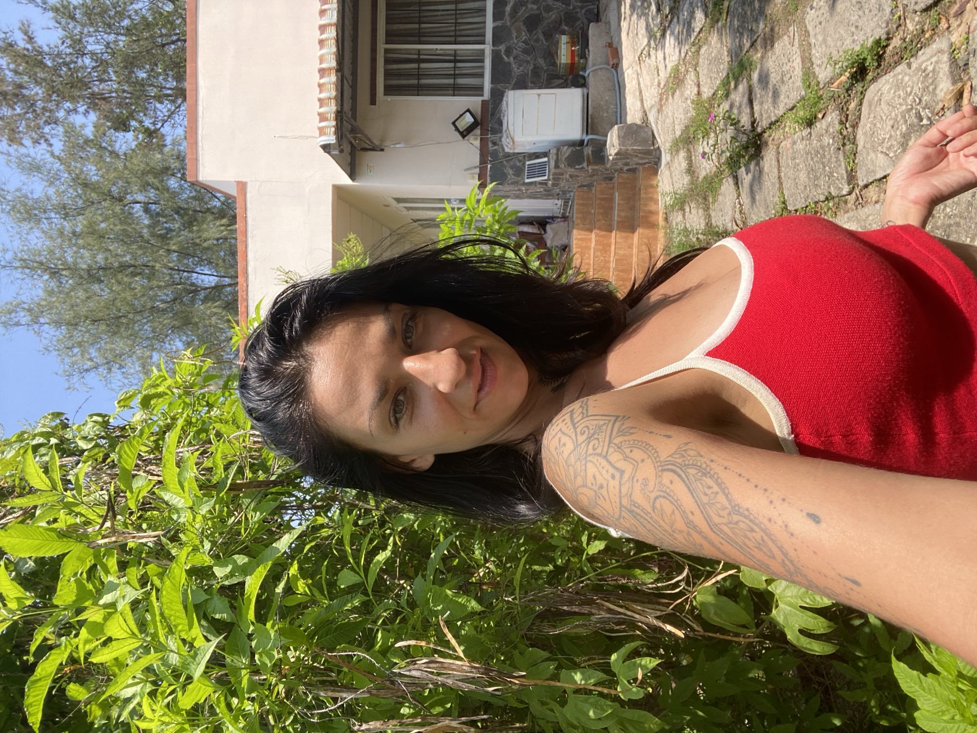 A woman is taking a selfie outdoors in what appears to be the city of Mui Ne, Vietnam. She has long, dark hair and is wearing a red tank top with white trim. She has a tattoo on her left arm. She is standing in front of lush green foliage and a building with a red-tiled roof. The ground is paved with stones and there are some plants growing between them. The sky is clear and blue. The overall impression is one of a sunny, tropical day.